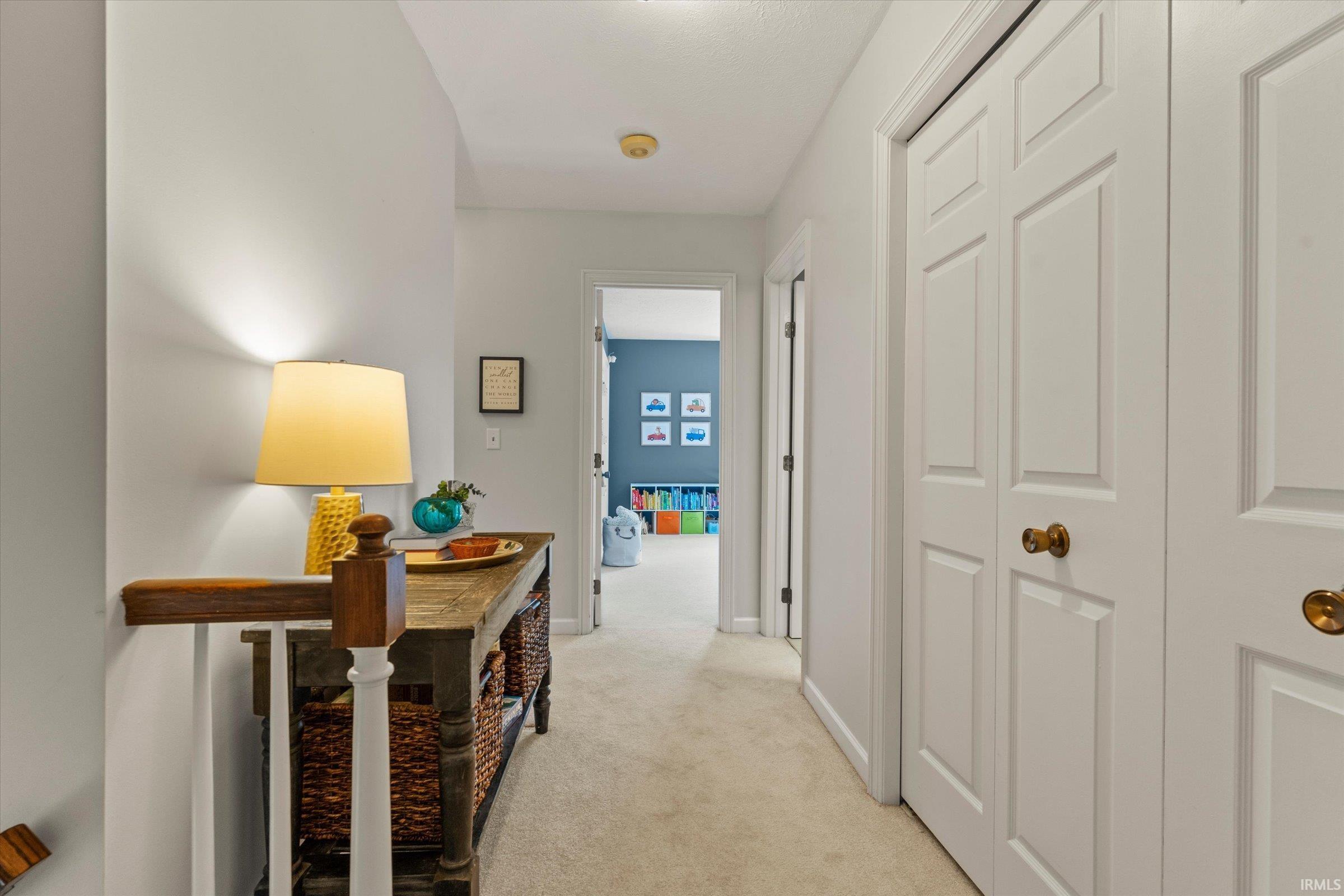 Hallway featuring light neutral wall paint, light-colored carpet flooring, white paneled doors with brass hardware, and a wood stair railing