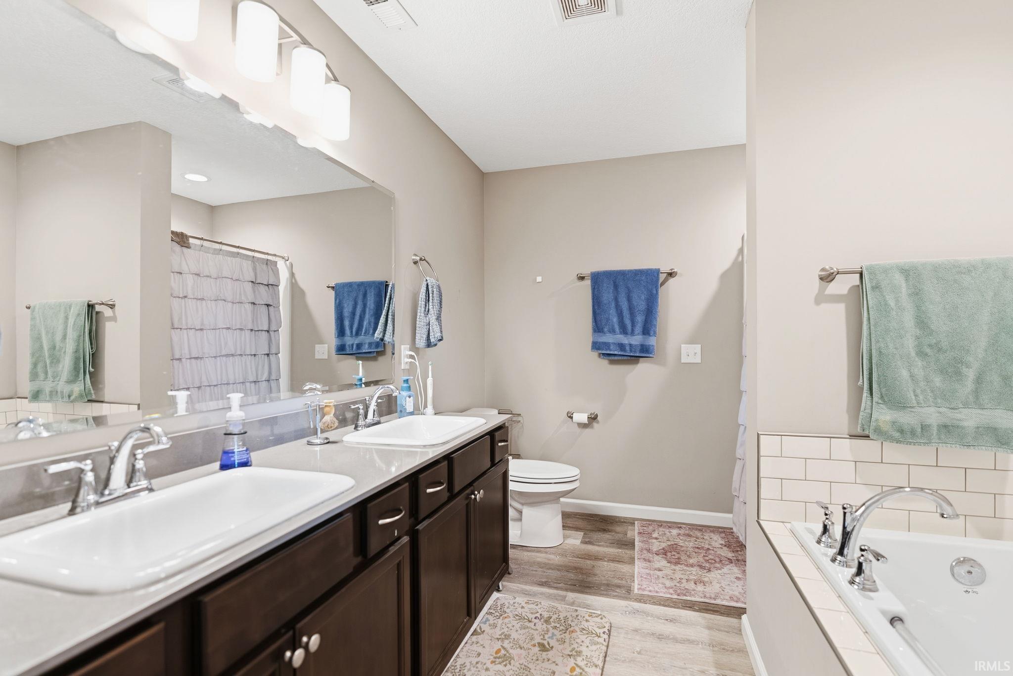Bathroom featuring a double vanity with ample storage, a large mirror, dual sinks with chrome fixtures, and a soaking tub with subway tile surround