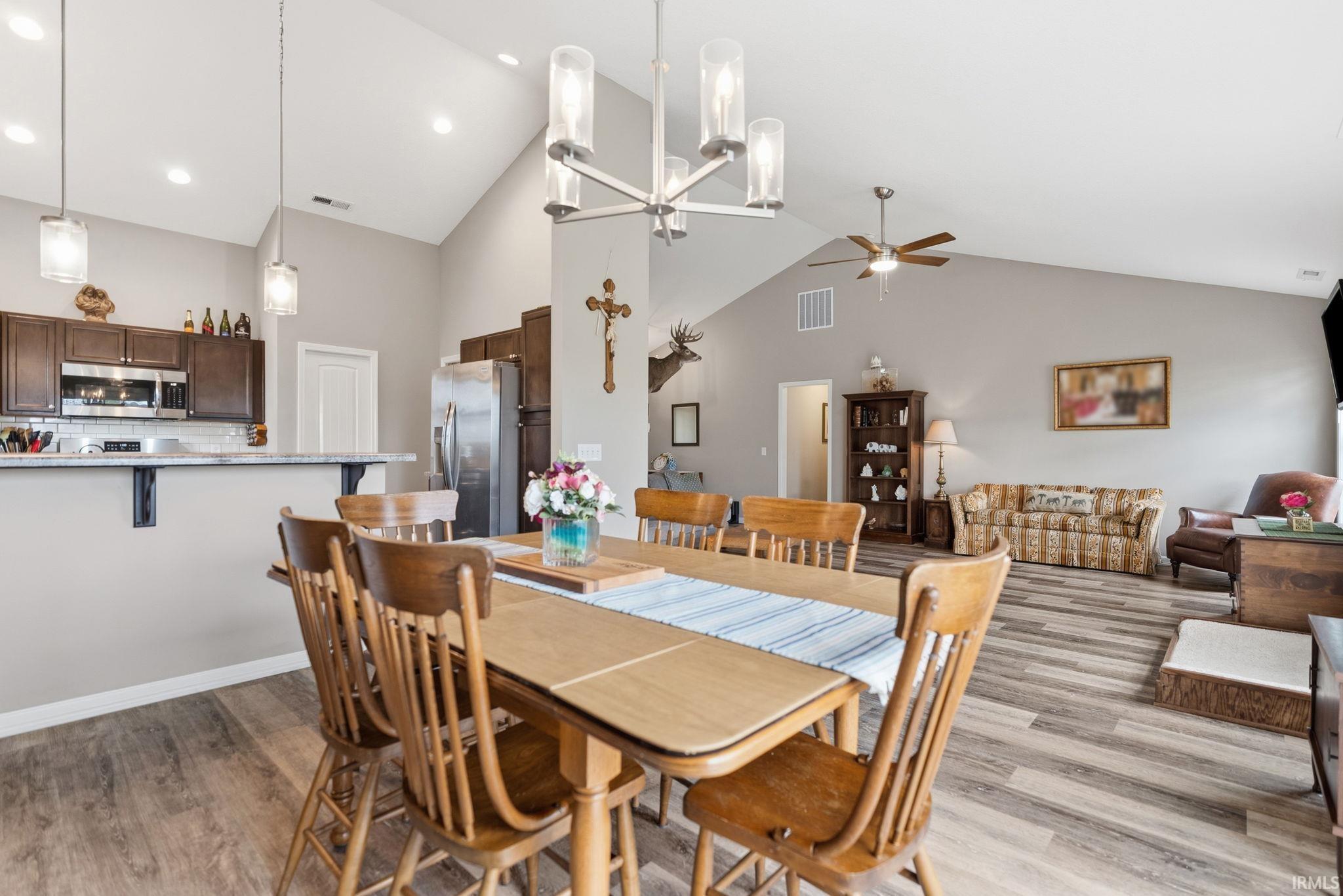 Open-concept living space featuring vaulted ceilings, wood-finish flooring, a kitchen with dark cabinetry and stainless steel appliances, a breakfast bar, and a dining area with pendant lighting