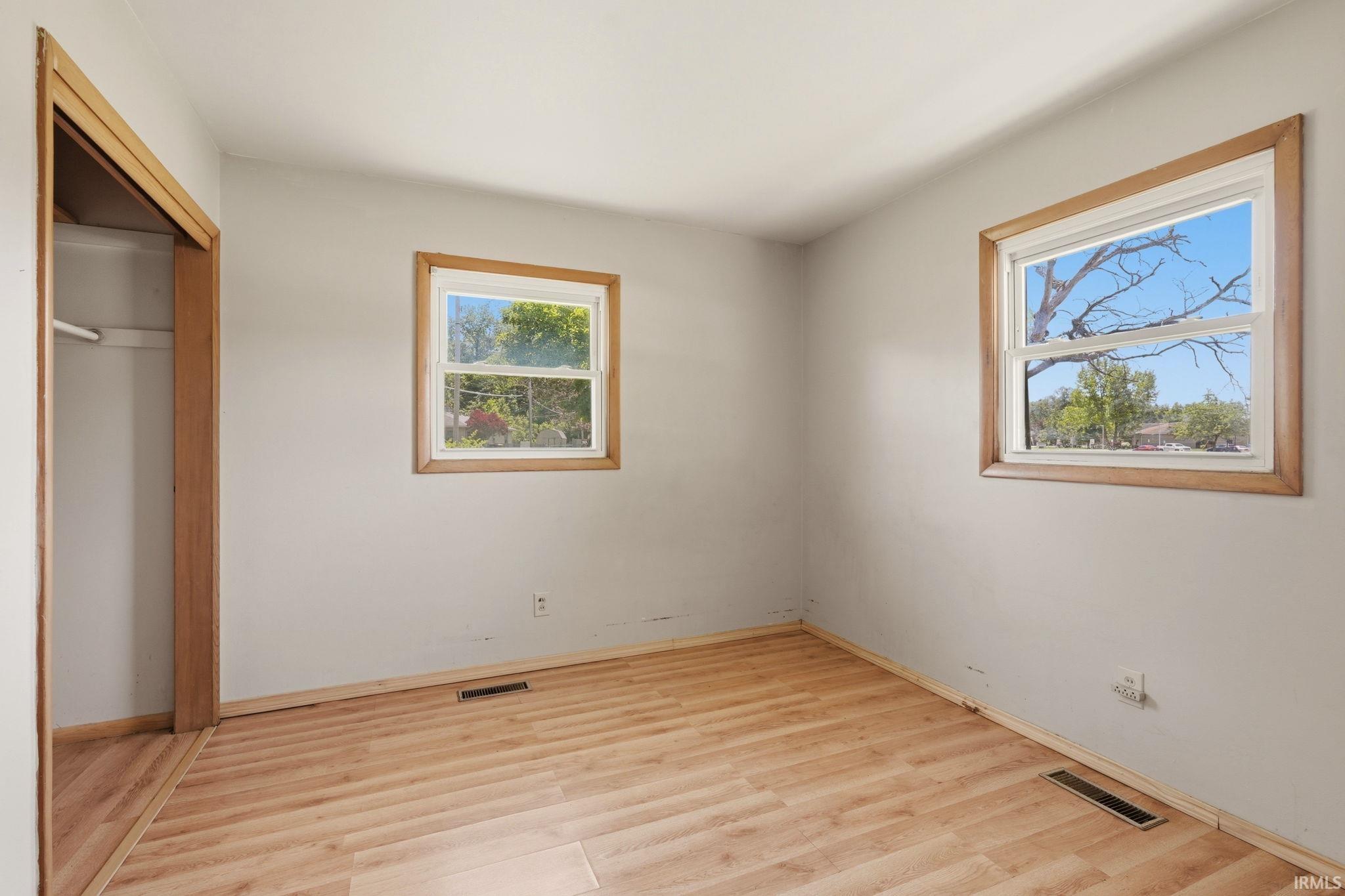 Room featuring light-toned wood-finish flooring, two windows with wood trim, and a wardrobe opening with a single shelf and hanging rod
