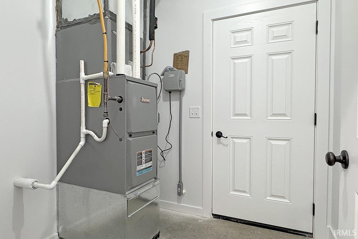 Utility room featuring a furnace with visible piping, a white four-panel interior door with a black handle, and concrete flooring