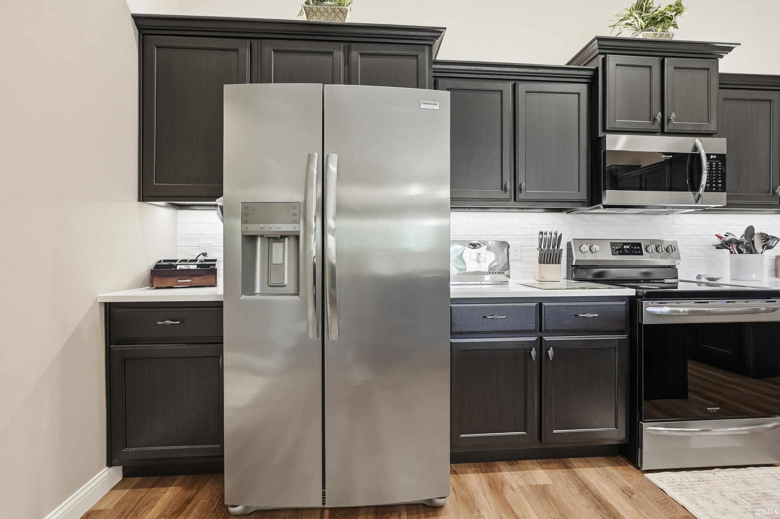 Kitchen featuring stainless steel appliances, dark wood cabinetry, white countertops, a tiled backsplash, and wood-finish flooring