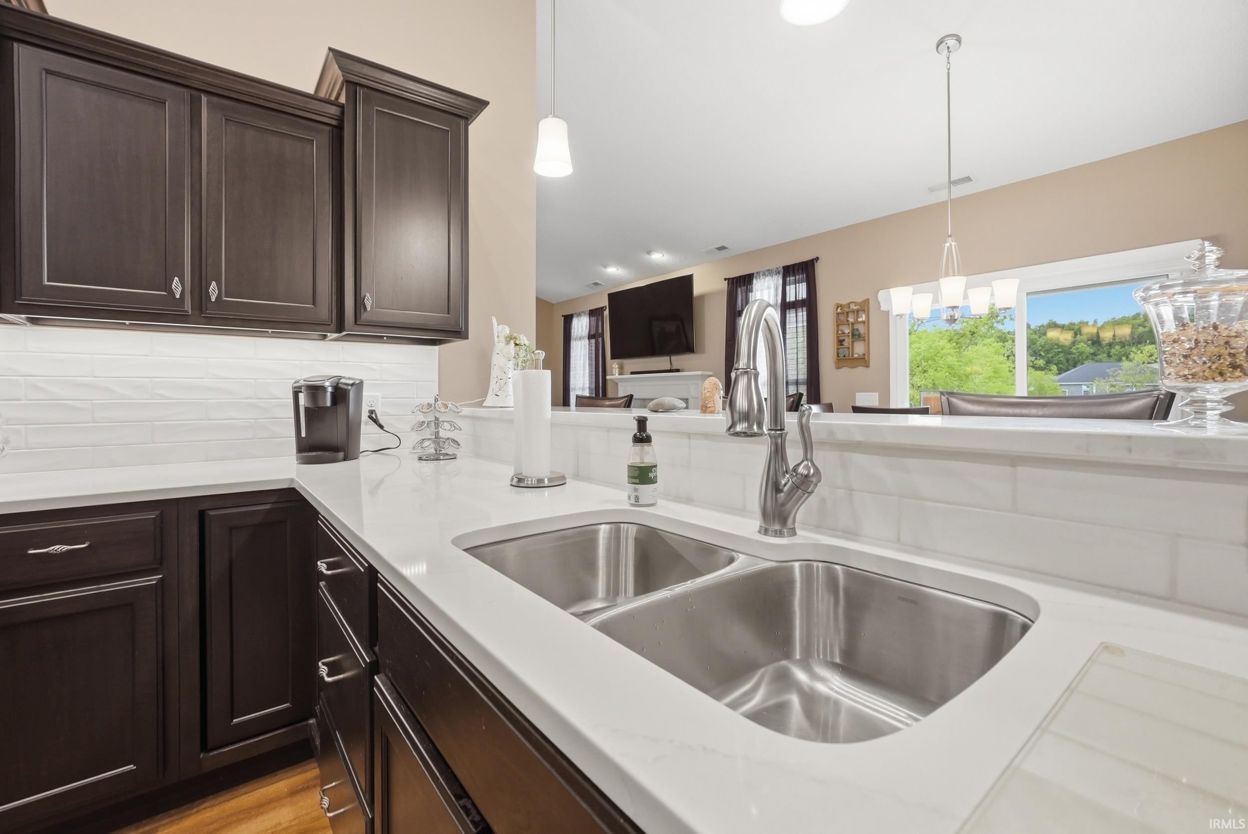 Kitchen featuring a stainless steel double-basin sink, gooseneck faucet, light-toned countertops, dark wood-finish cabinetry with silver hardware, and a white subway tile backsplash