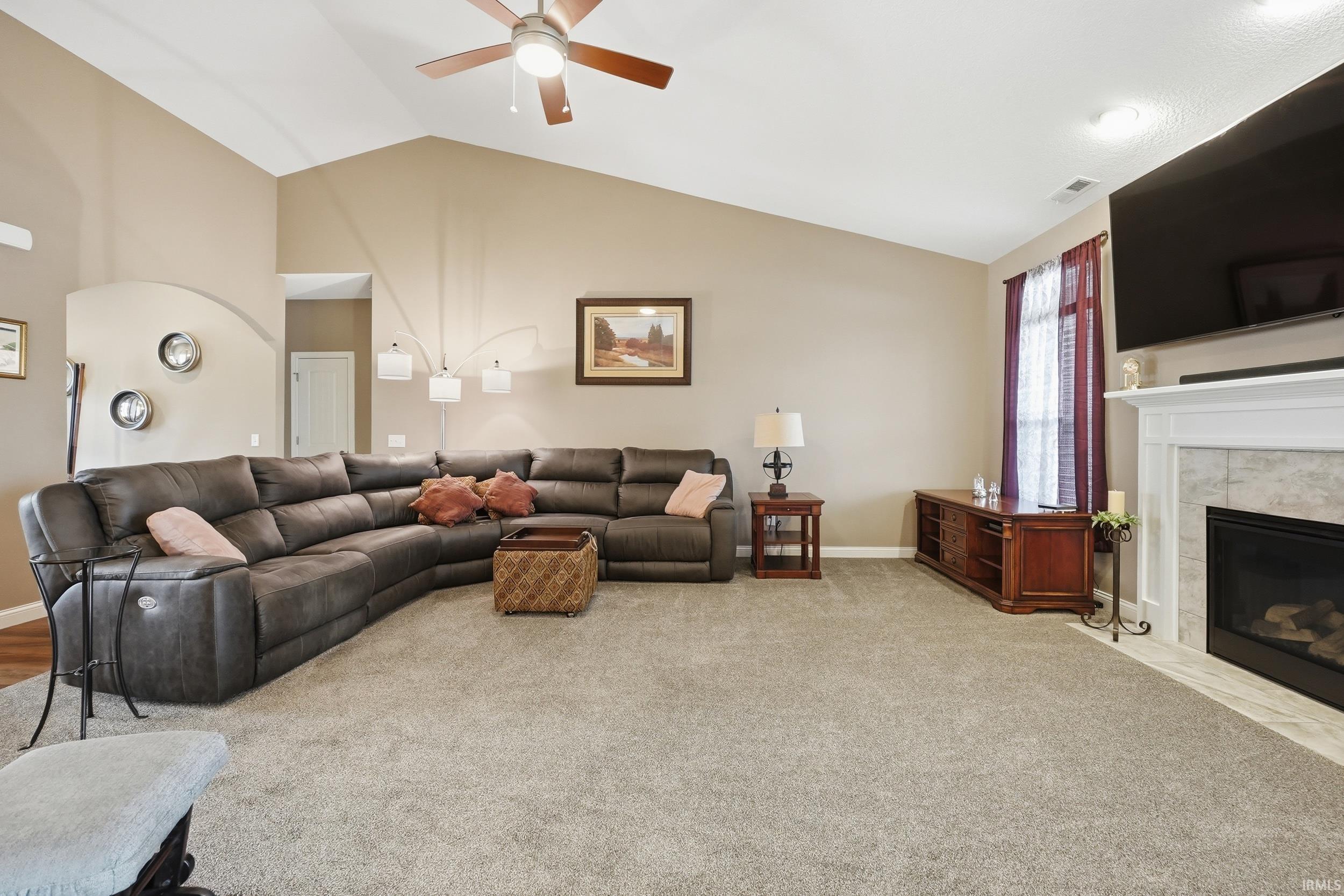 Vaulted ceiling living area featuring a ceiling fan with light fixture, carpeted flooring, and a fireplace with tile surround and white mantel