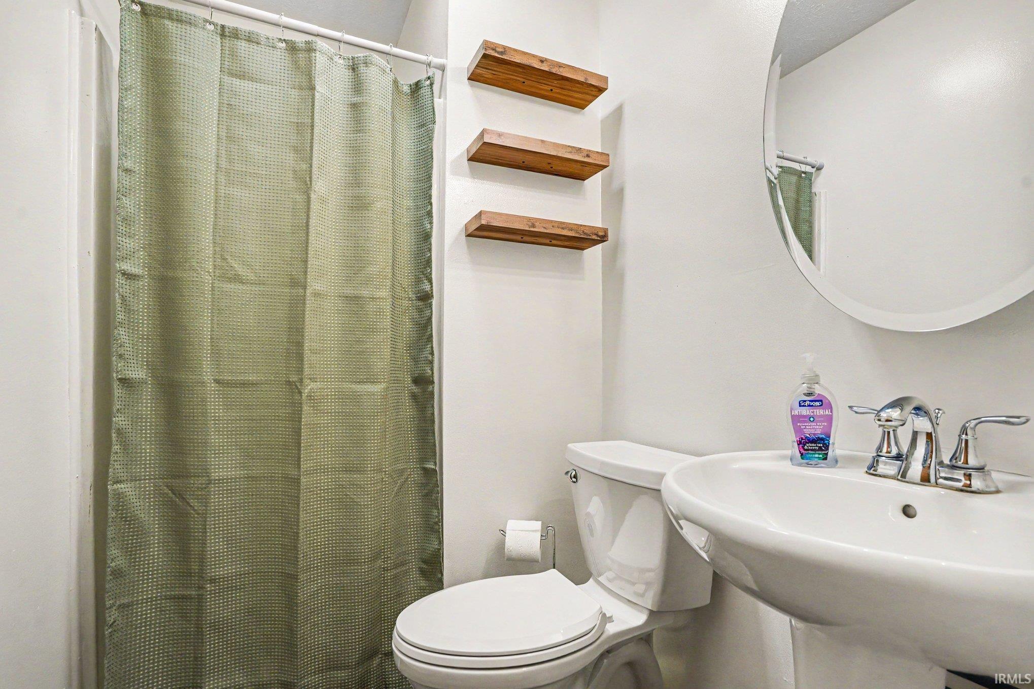 Bathroom featuring a pedestal sink with chrome fixtures, a round mirror, and three wall-mounted wood shelves