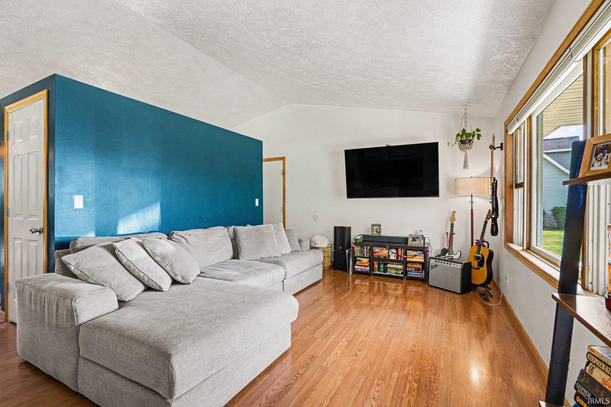 Living area featuring wood-finish flooring, a vaulted ceiling, multiple windows with wood trim, and a prominent accent wall