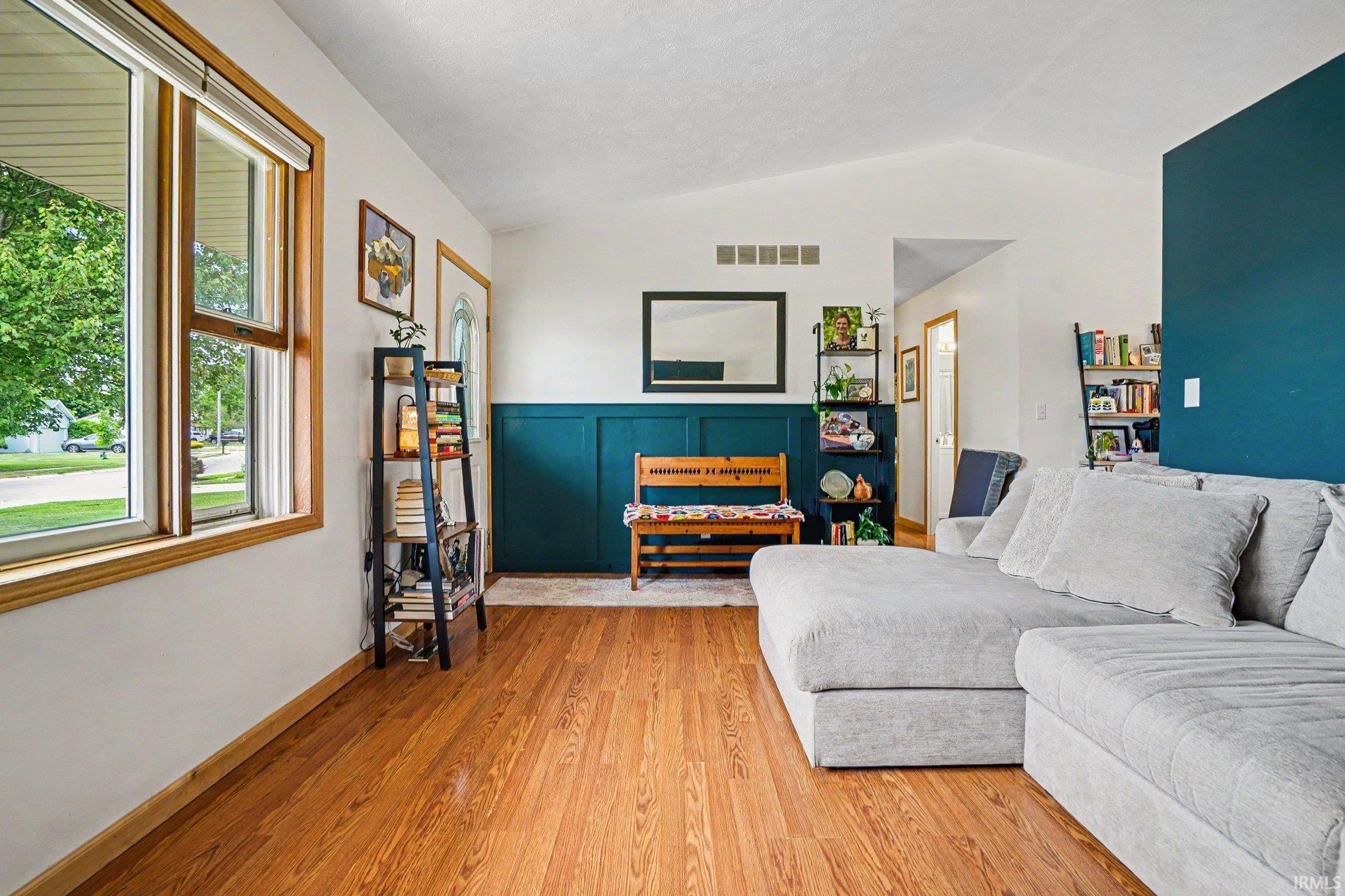 Vaulted ceiling living space featuring wood-finish flooring, a large window with wood trim, and accent wainscoting