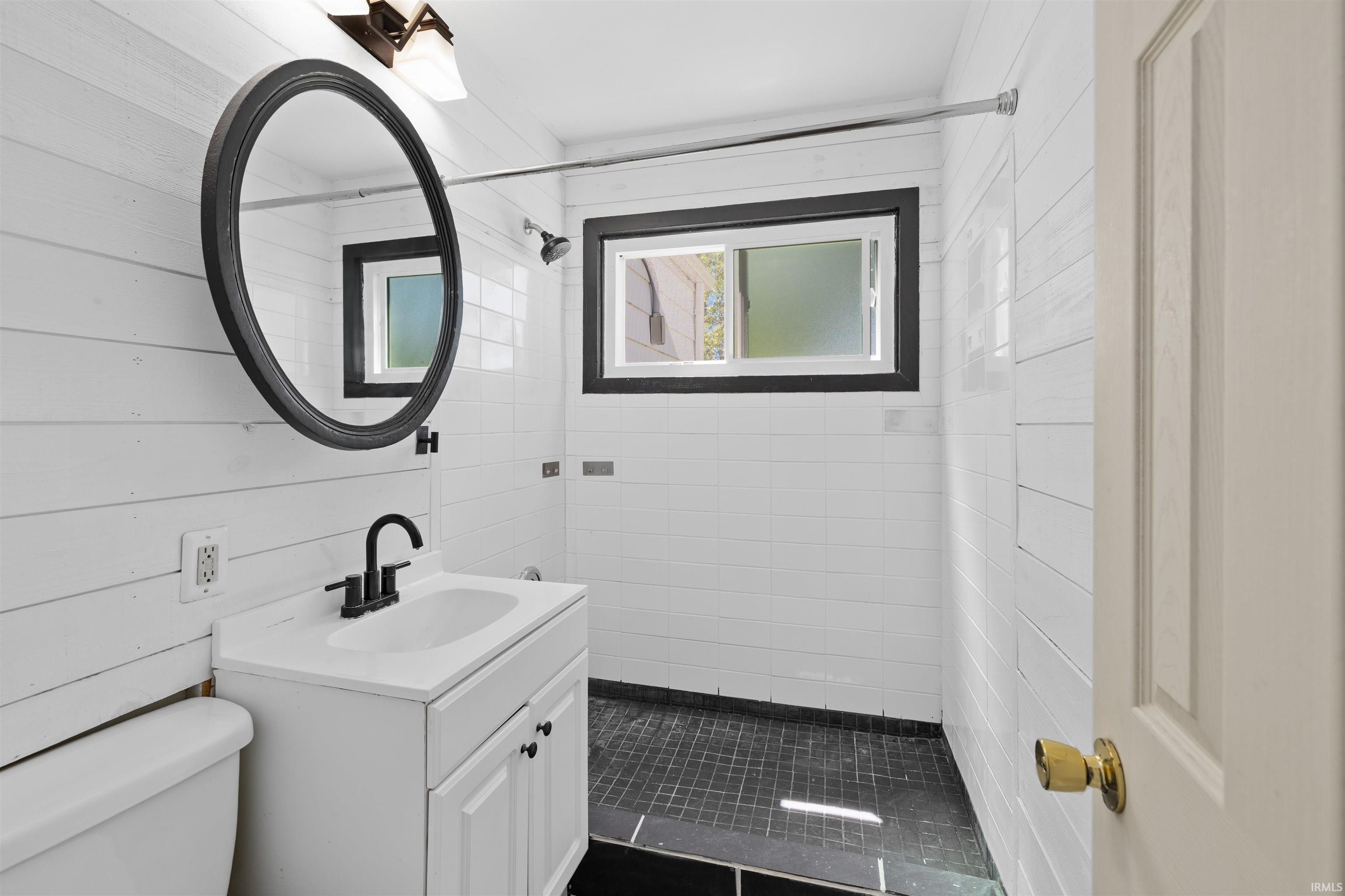 Bathroom featuring shiplap wall paneling, white subway tile shower surround, black mosaic tile shower floor, black-framed mirror, and a white vanity with matte black hardware
