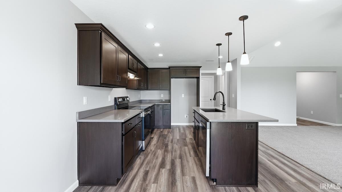 Kitchen featuring dark wood-finish cabinetry, light-toned countertops, a central island with a sink and pendant lighting, and wood-finish flooring