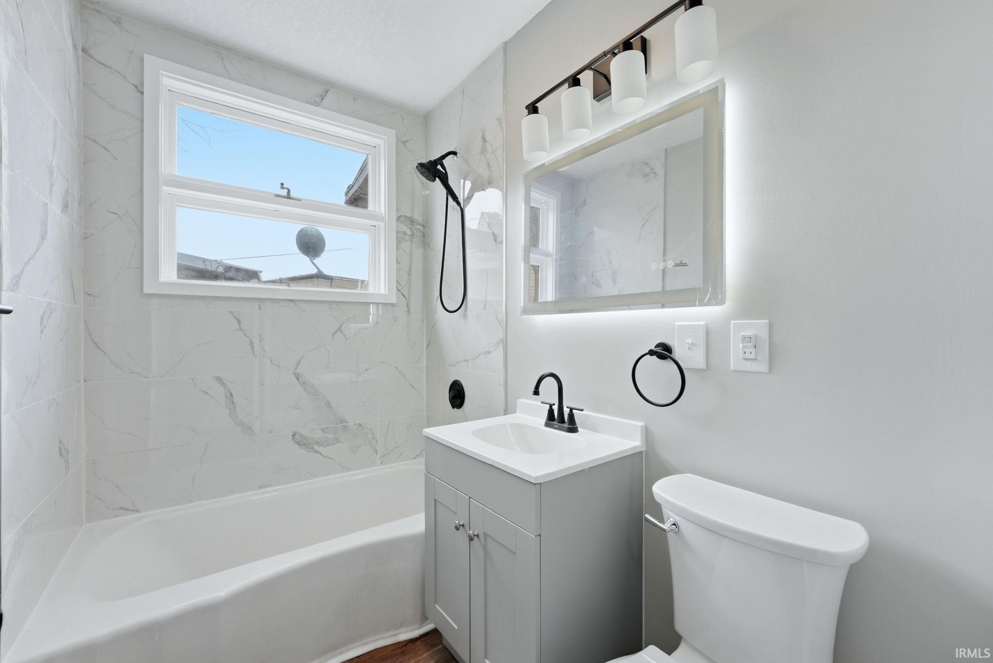 Contemporary bathroom featuring a white porcelain bathtub, a vanity with a light gray cabinet and white countertop, an illuminated mirror, and matte black fixtures
