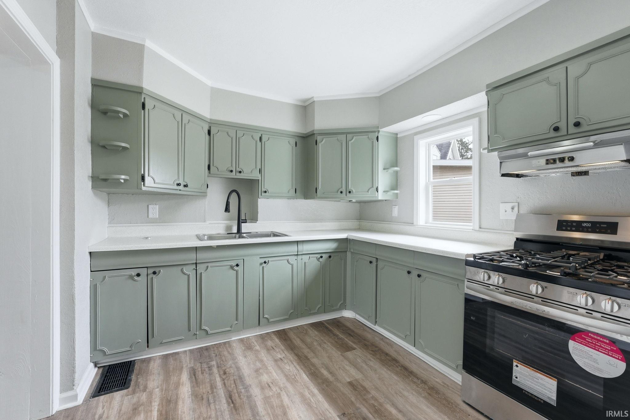 Kitchen featuring sage green cabinetry, light-toned countertops, wood-finish flooring, a stainless steel range with a coordinating range hood, and a window for natural light