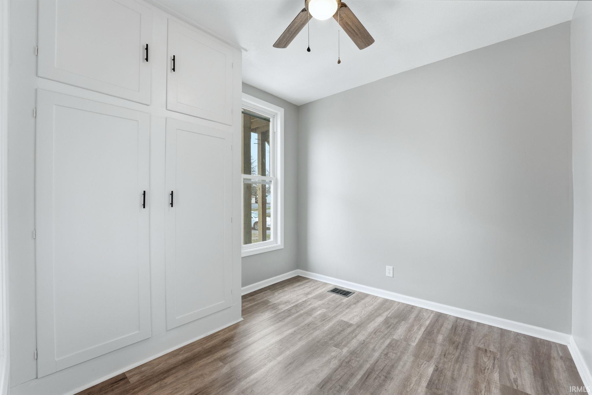 Interior room featuring built-in white cabinetry, wood-finish flooring, a ceiling fan with integrated lighting, and a double-hung window