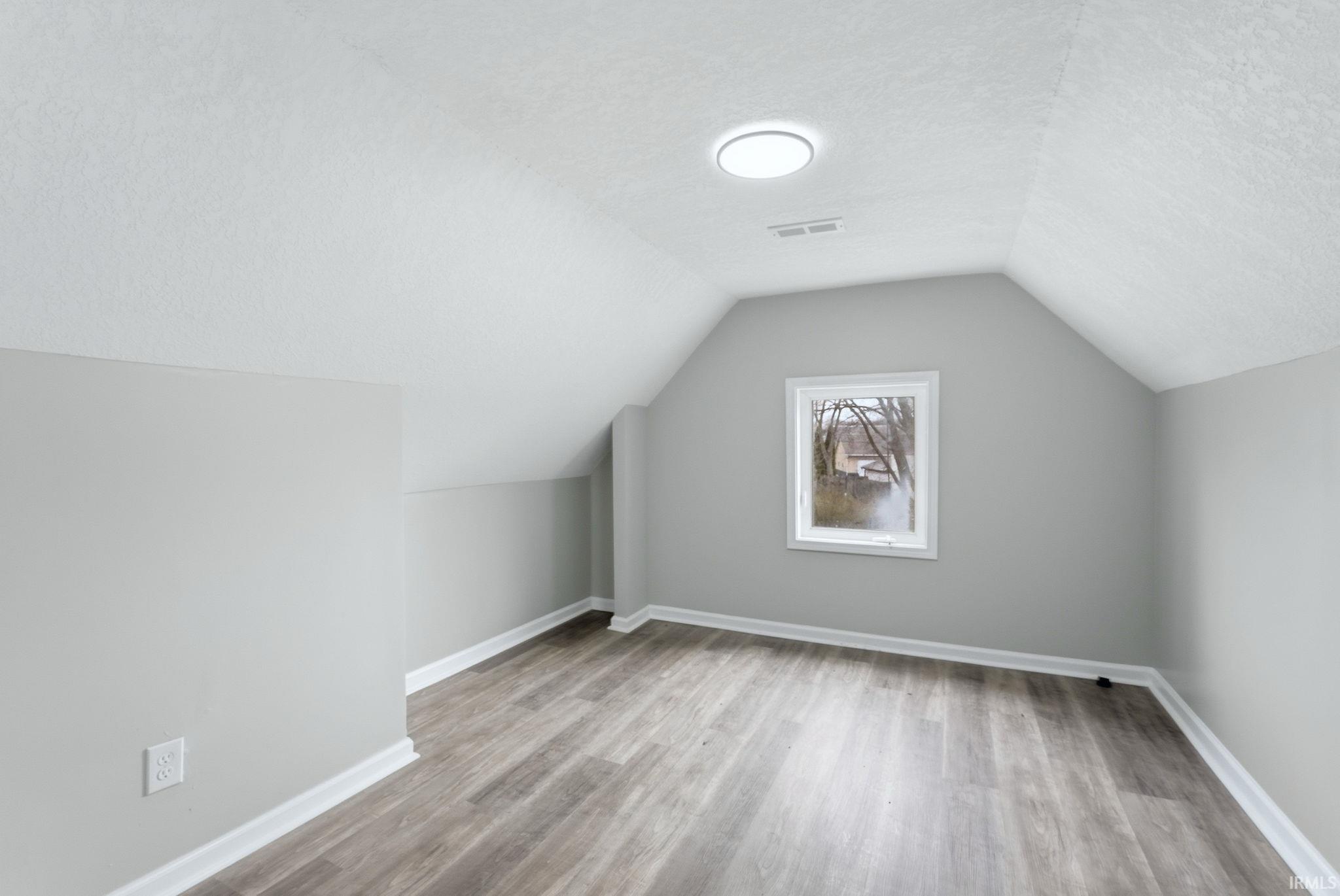 Attic room featuring wood-finish flooring, light gray walls, white trim, and a prominent window