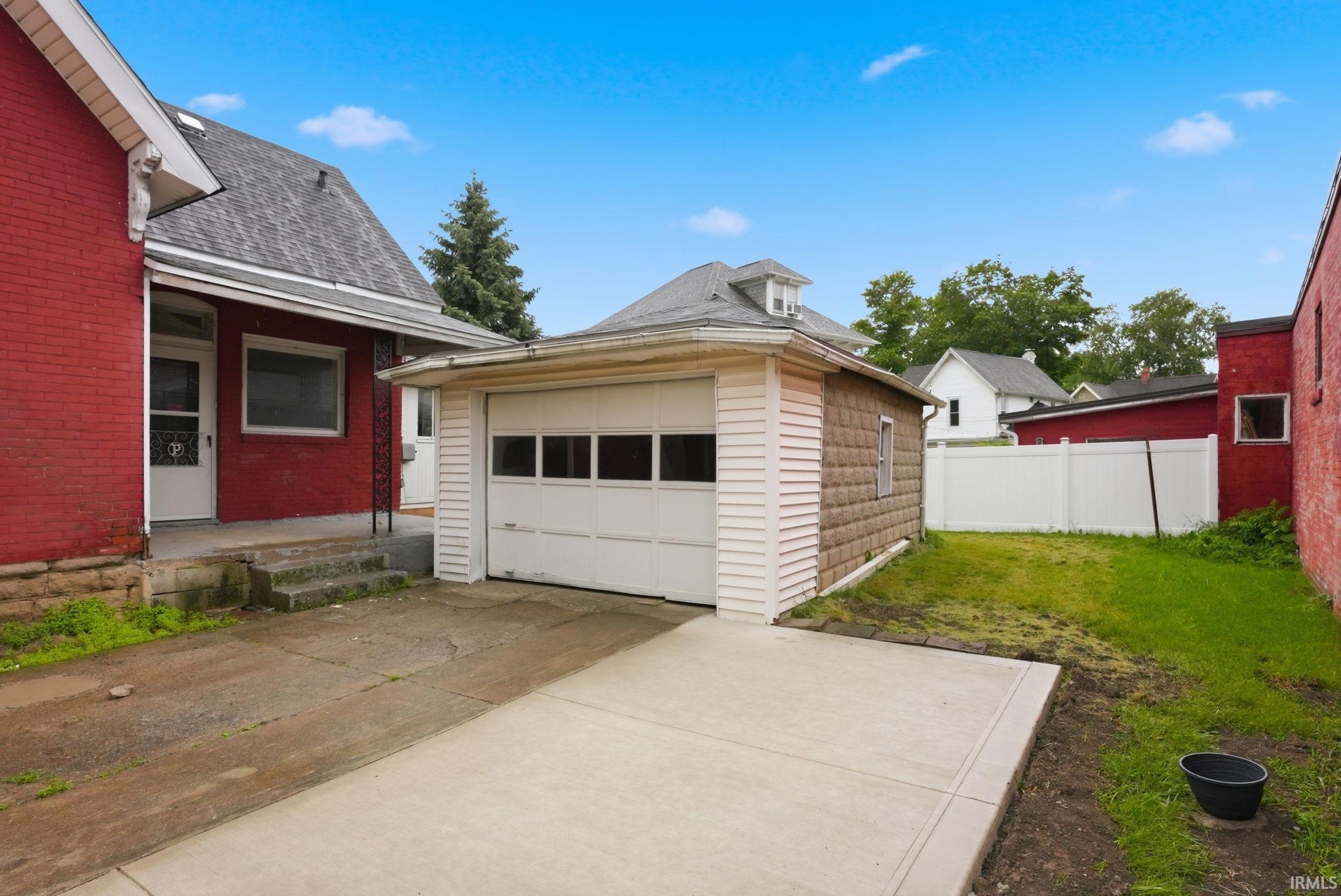 Detached garage with a paneled door, a newly poured concrete driveway, a red brick exterior, a white privacy fence, and a grassy yard space