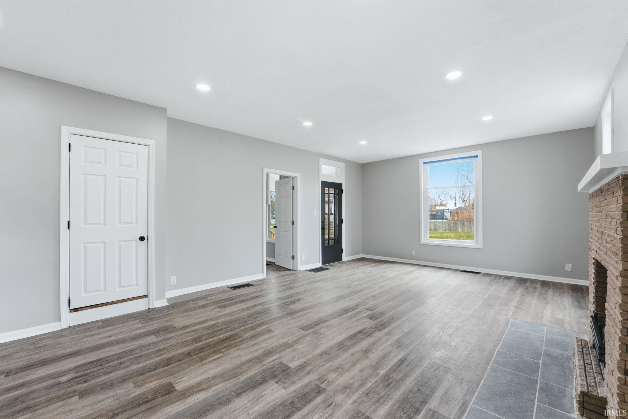 Spacious room featuring wood-finish flooring, light grey walls, recessed lighting, a fireplace with a brick surround and stone hearth, and a large window providing natural light