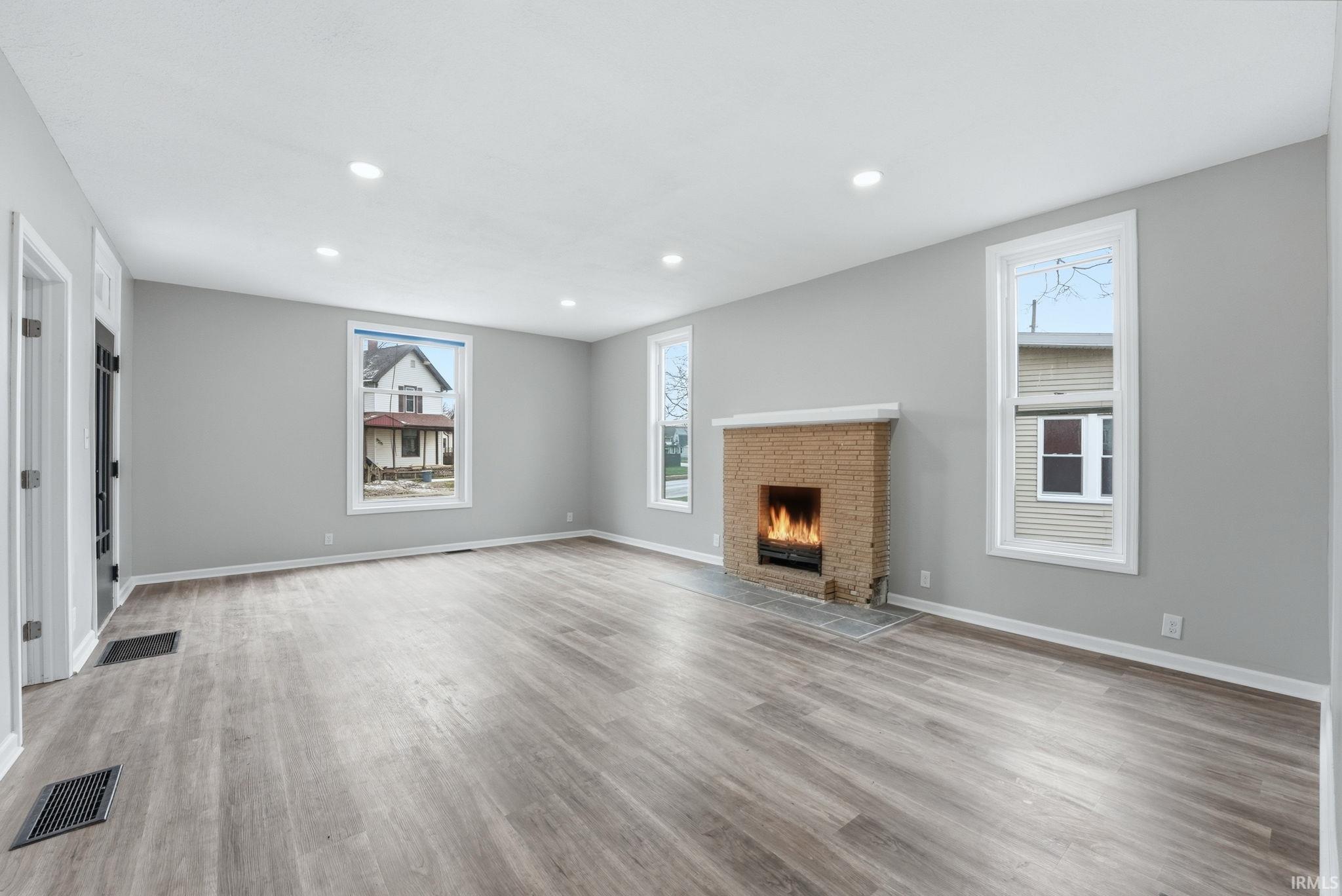 Spacious living area featuring light wood-finish flooring, a brick fireplace with mantel, recessed ceiling lighting, multiple windows, and a neutral wall color