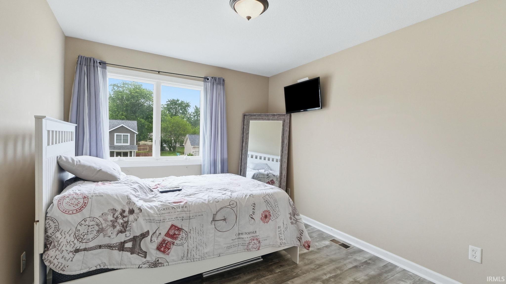 Neutral-toned bedroom featuring wood-finish flooring, a white ceiling, and a large window with white trim and light-colored curtains