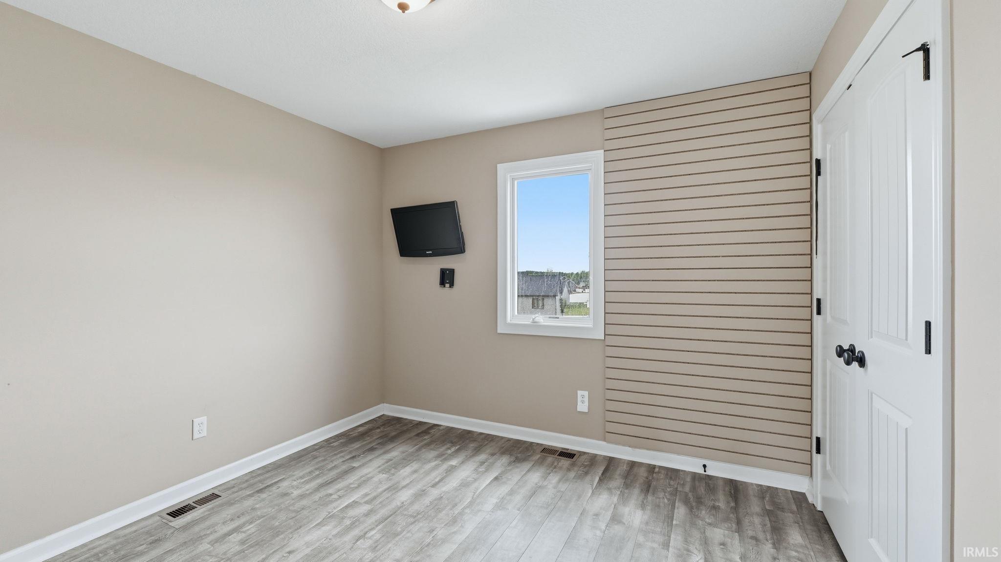 Bedroom featuring a window with exterior views, wood-finish flooring, a white paneled door with black hardware, and slat wall paneling