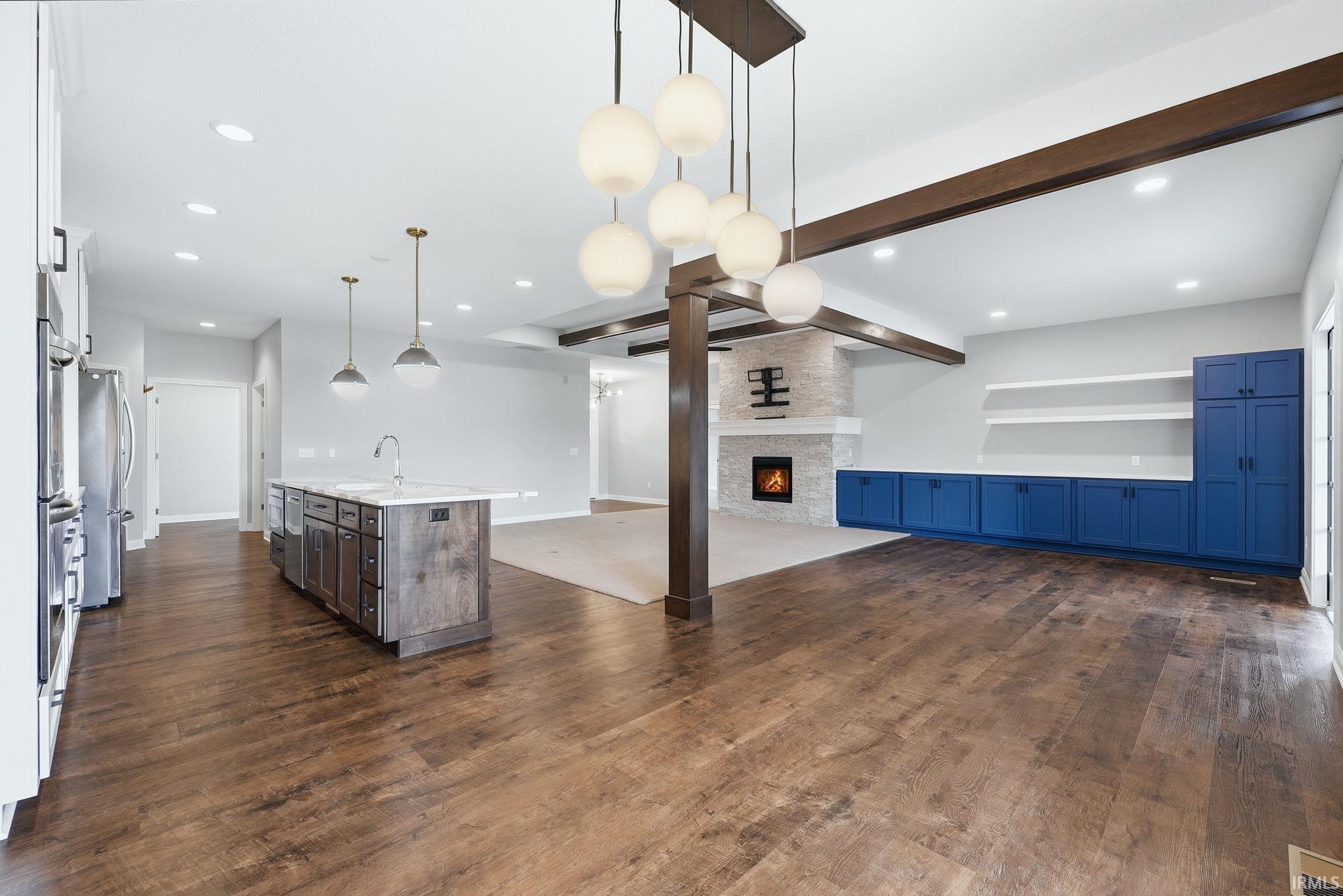 Spacious open-concept interior featuring wood-finish flooring, a kitchen island with a built-in sink, a stone-clad fireplace, built-in blue cabinetry, and exposed ceiling beams