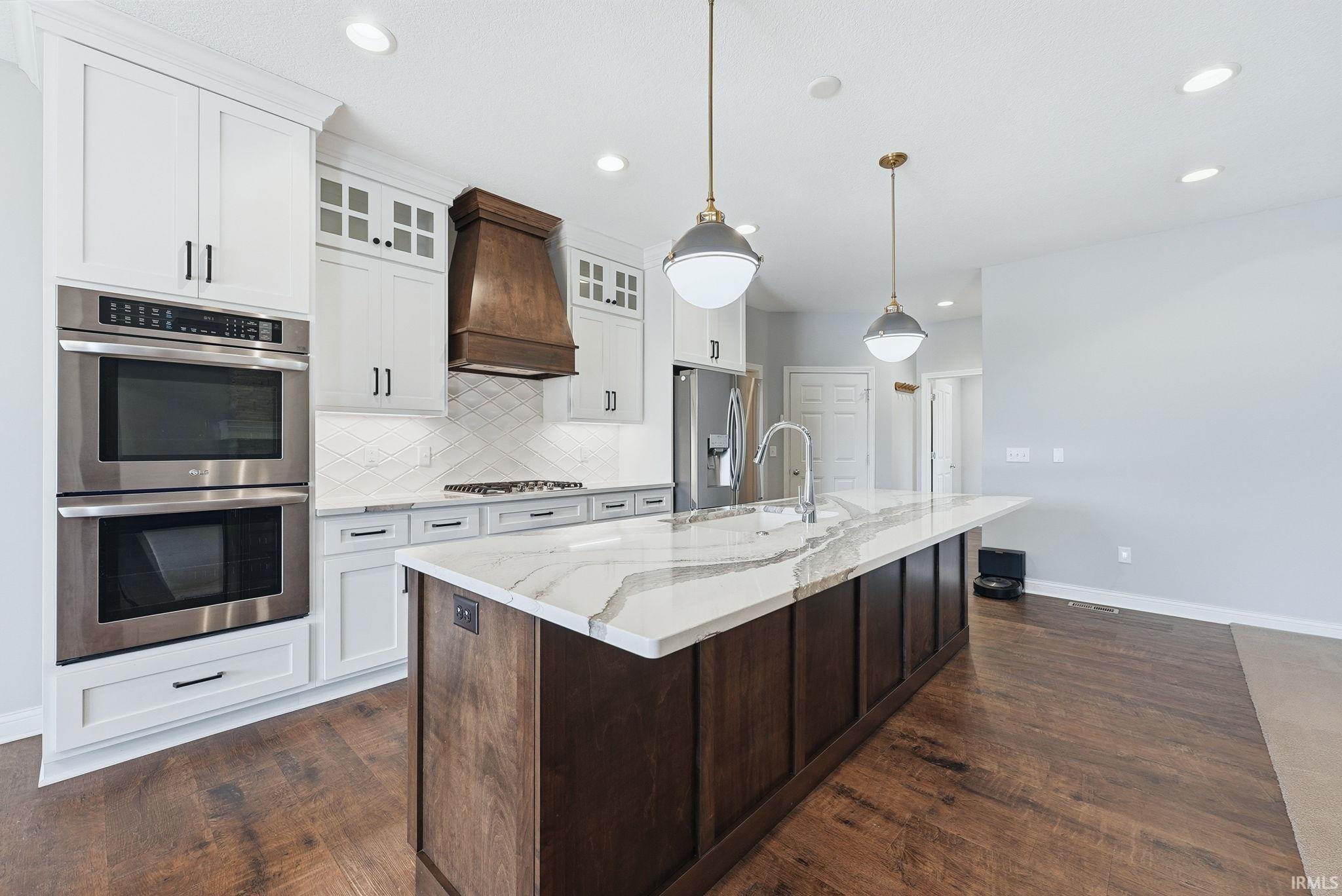 Kitchen featuring a large center island with a stone countertop, stainless steel appliances including a double oven, wood-finish flooring, white cabinetry, and a decorative wood range hood