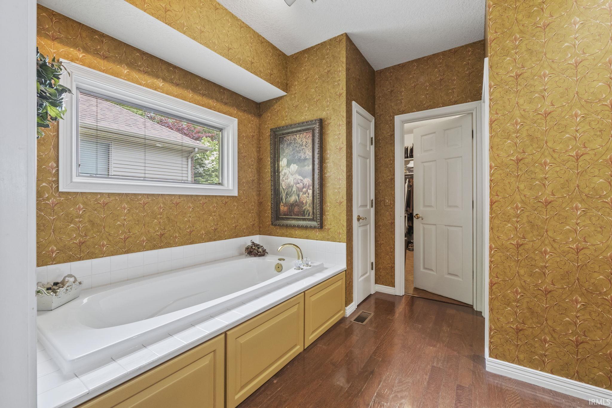 Soaking tub with white tile surround, recessed storage cabinets, and wood-finish flooring