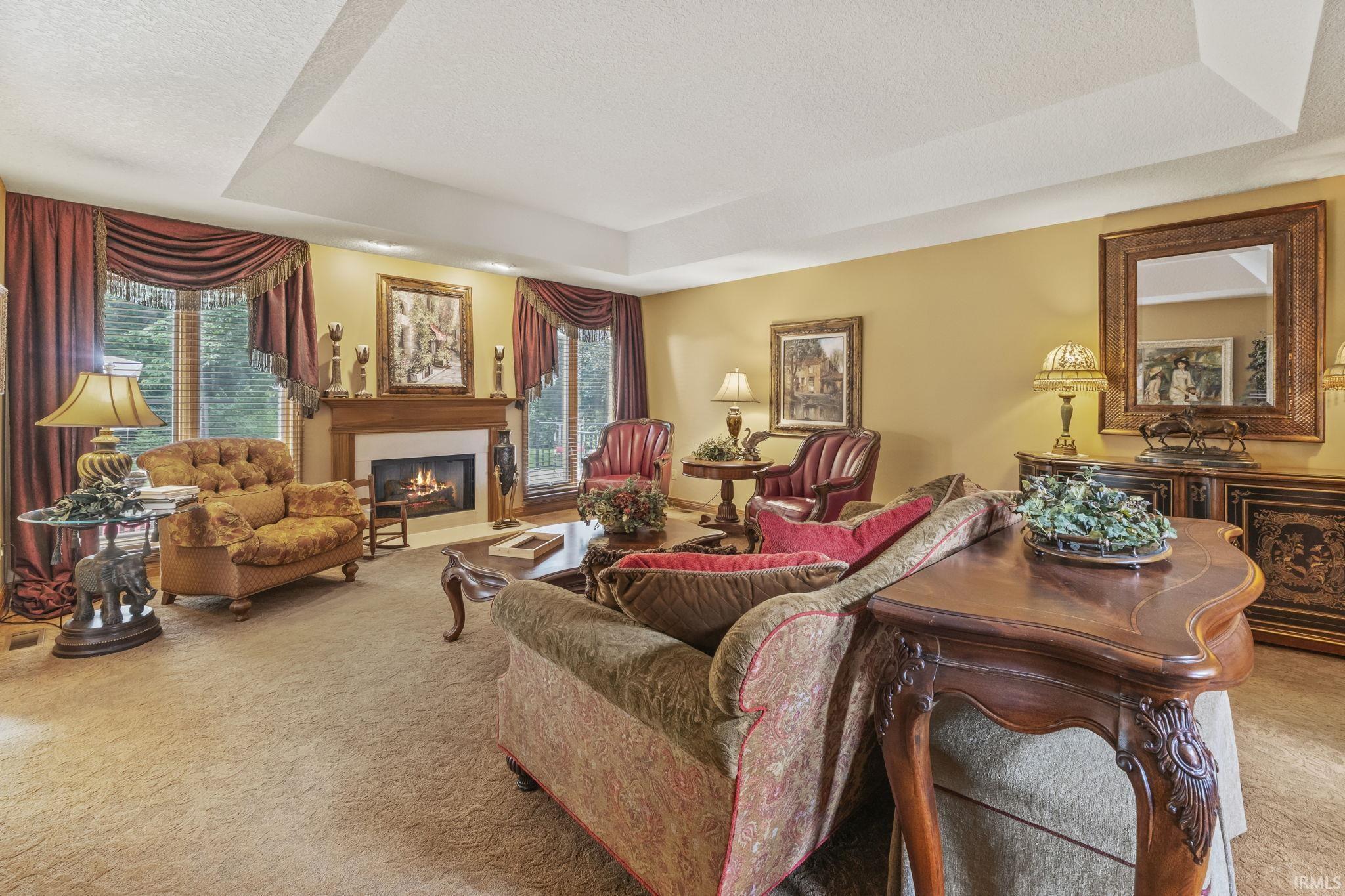 Spacious living area featuring a tray ceiling, a fireplace with a white surround and wood mantel, large windows with decorative drapery, and textured carpet flooring