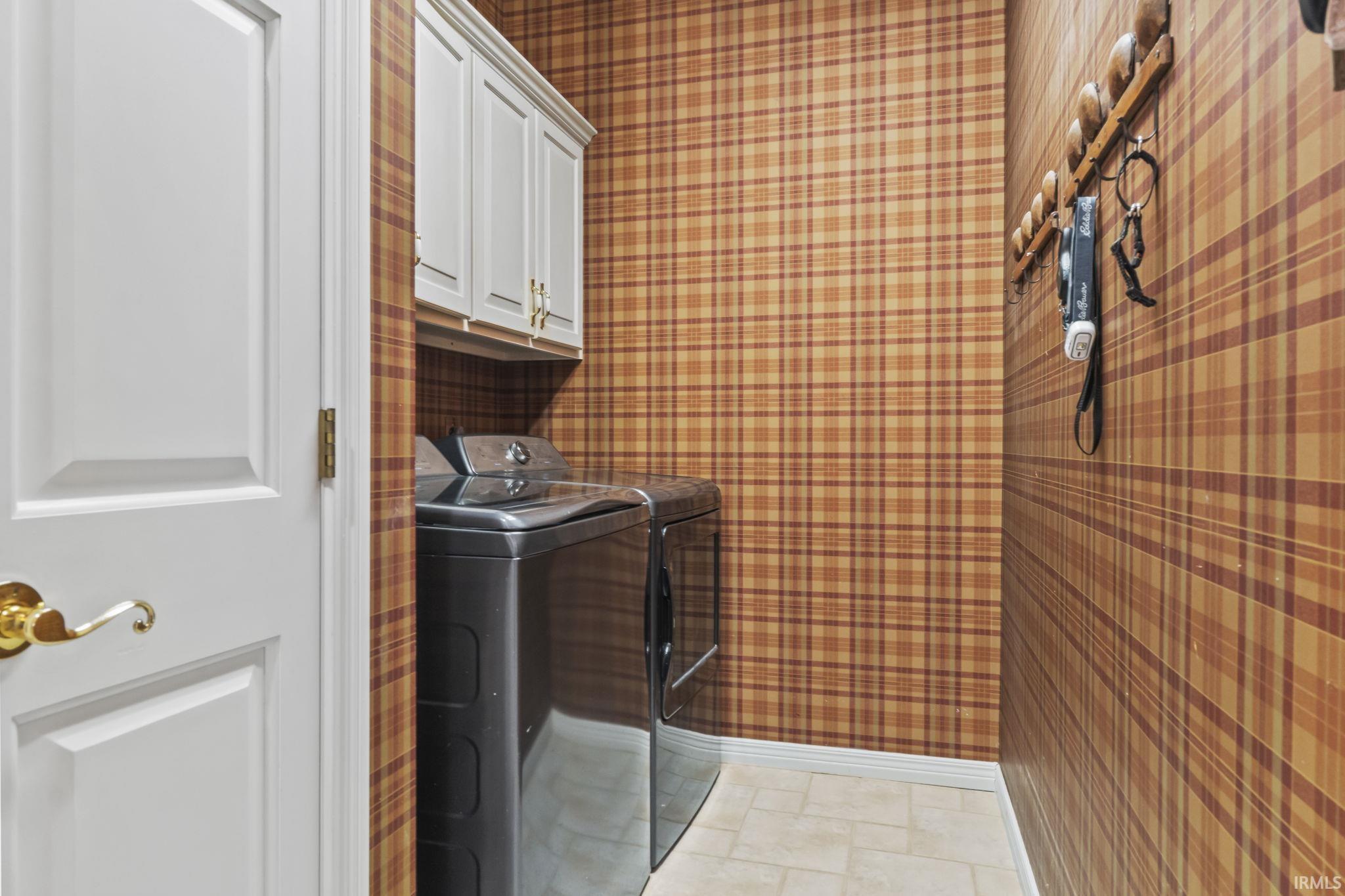 Laundry area featuring neutral plaid wall covering, white cabinetry with gold-tone hardware, and light-toned tile flooring