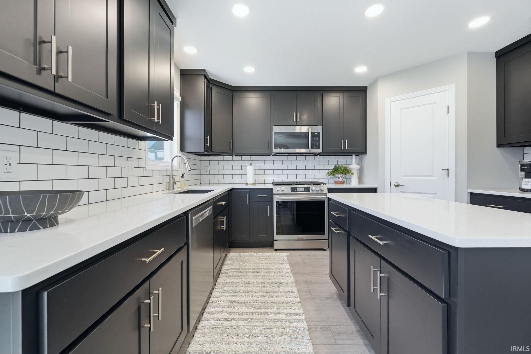 Modern kitchen featuring dark cabinetry with brushed nickel hardware, white subway tile backsplash, light countertops, stainless steel appliances, and recessed lighting