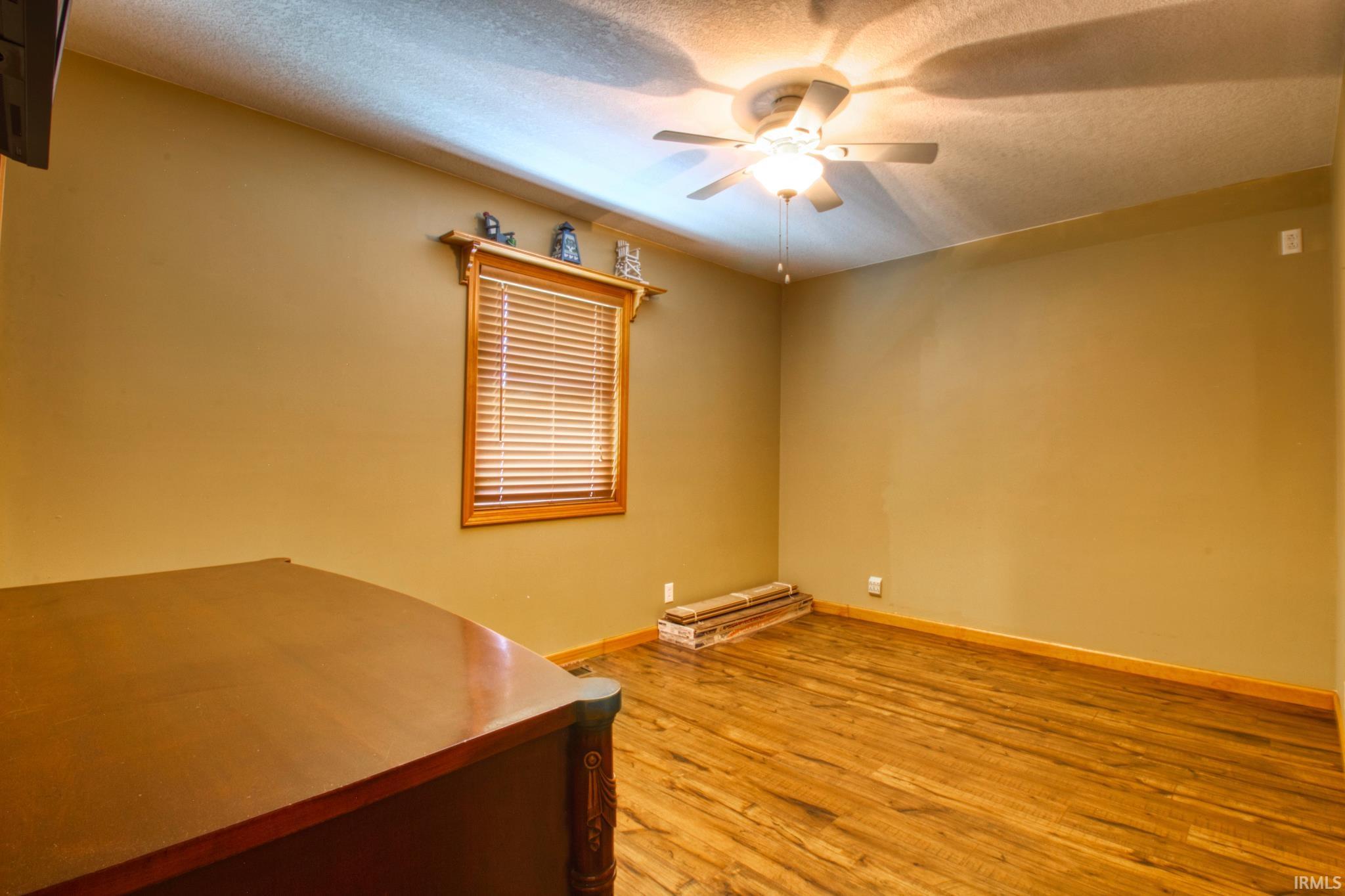 Interior room featuring wood-finish flooring, a window with wood blinds and trim, a decorative wall shelf, and a ceiling fan with integrated lighting