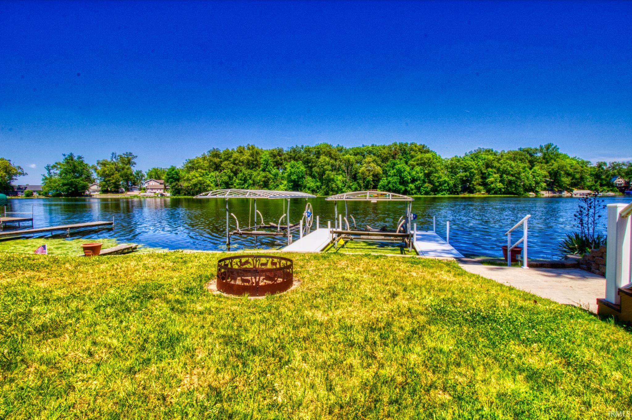 Expansive waterfront property featuring a manicured lawn, a built-in fire pit, a private dock, and two covered boat lifts on the lake