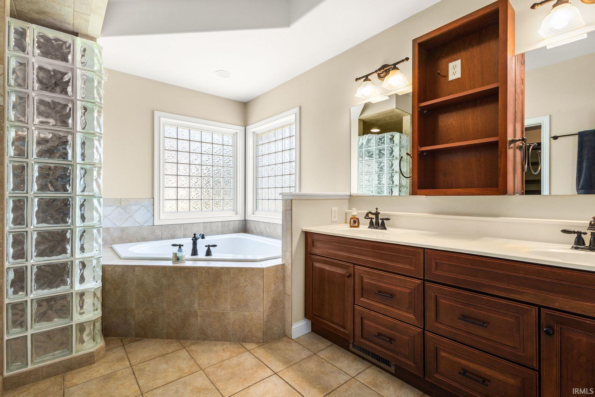 Bathroom featuring a corner soaking tub, glass block shower enclosure, dual basin vanity with dark wood cabinetry, and tiled flooring