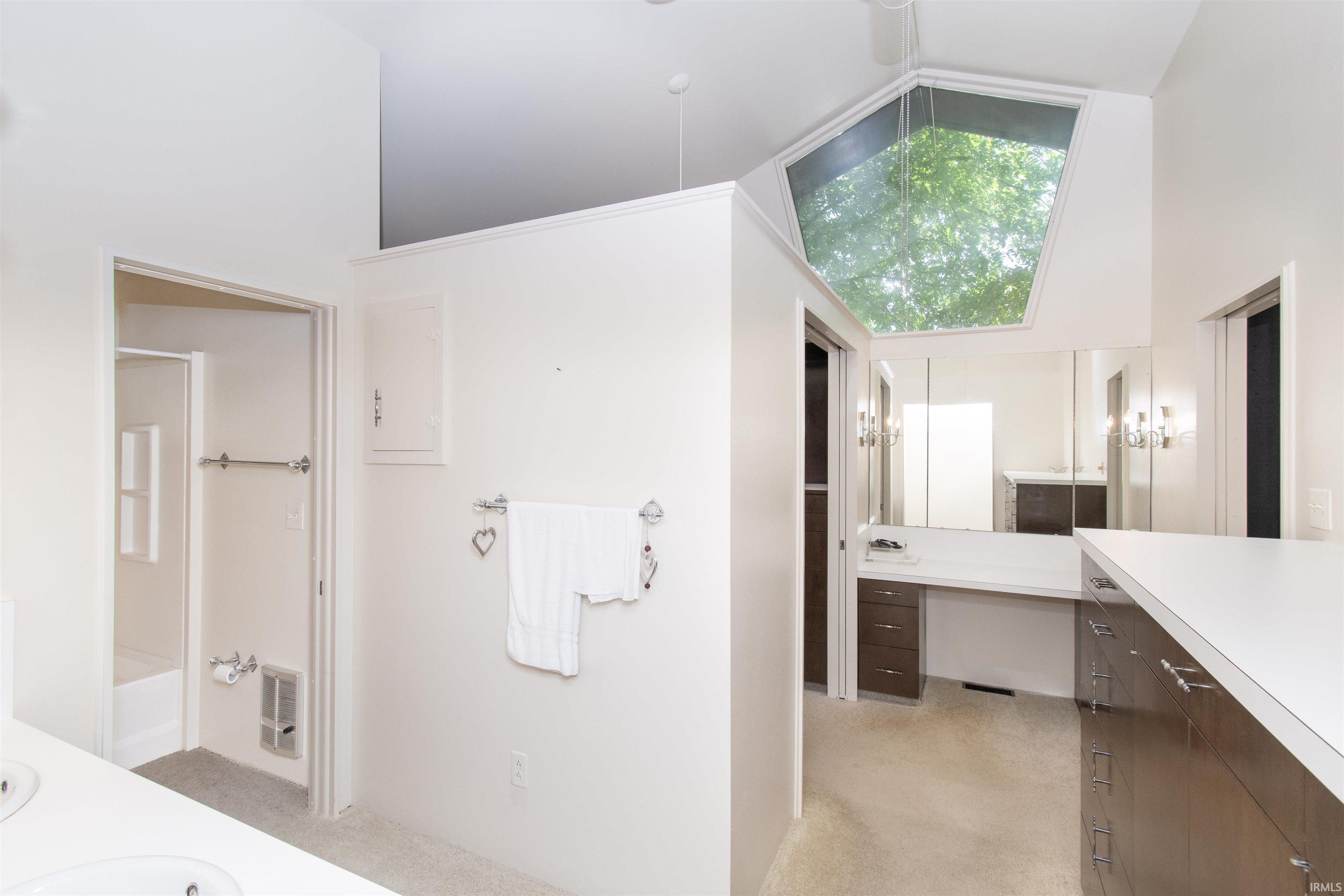 Vaulted ceiling with an angular skylight, vanity area with integrated mirrors and sconce lighting, white countertops, and dark wood-finish cabinetry