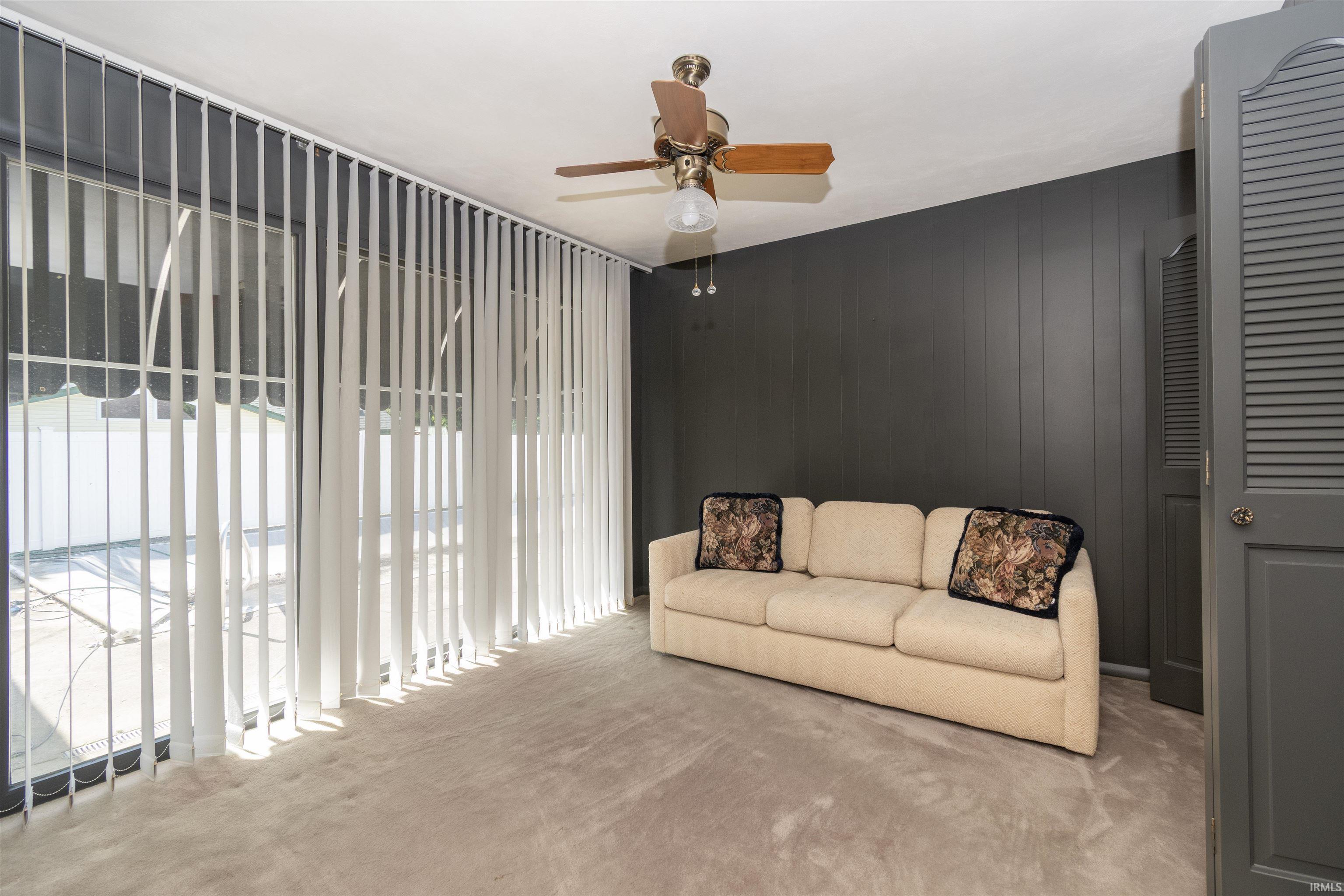 Spacious room featuring vertical blinds across a large window, dark paneled wall, light carpet flooring, ceiling fan with lighting, and a louvered bi-fold door