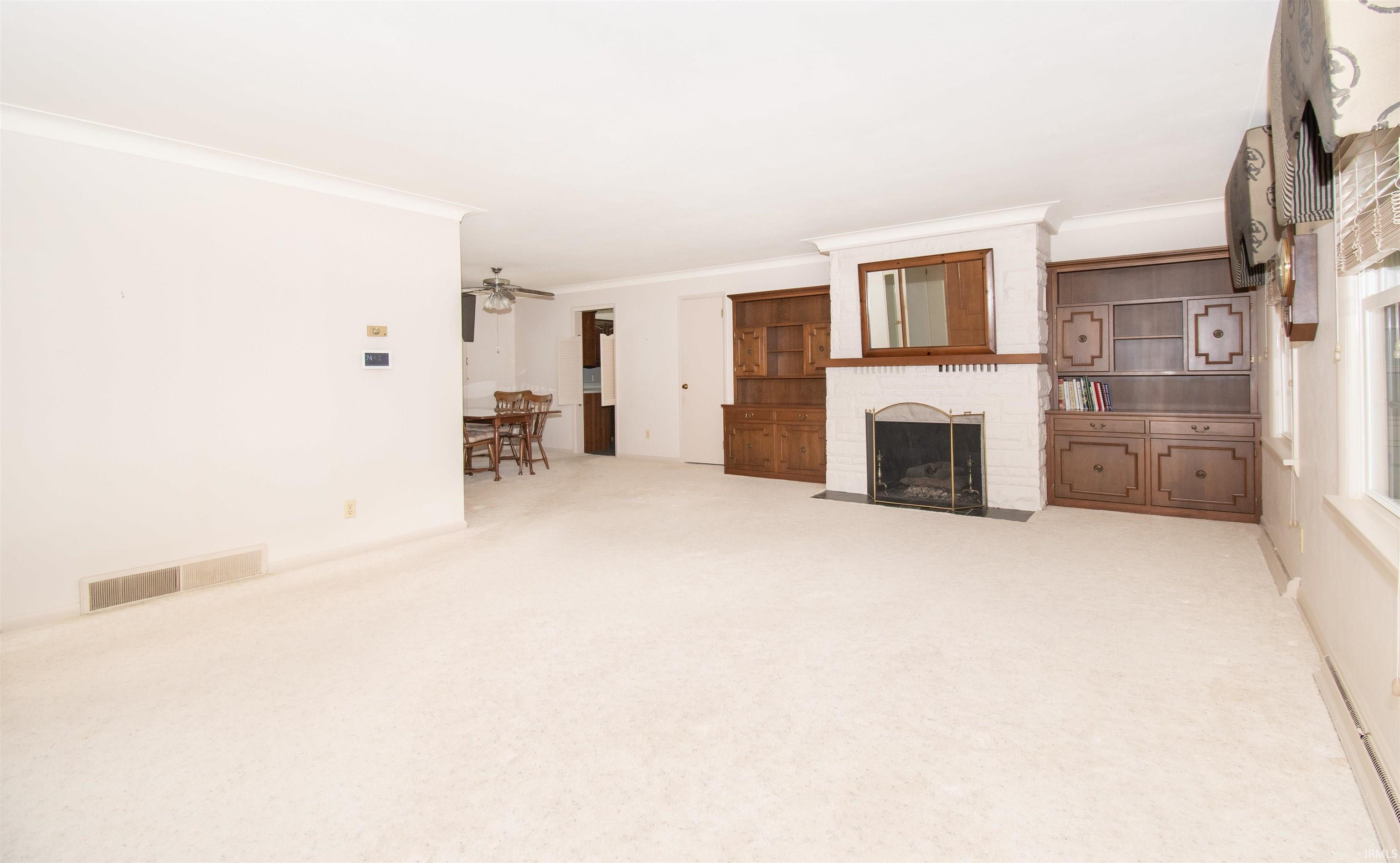Spacious living area featuring a white brick fireplace with a dark wood mantle and mirror, built-in dark wood cabinetry, white carpeting, and crown molding