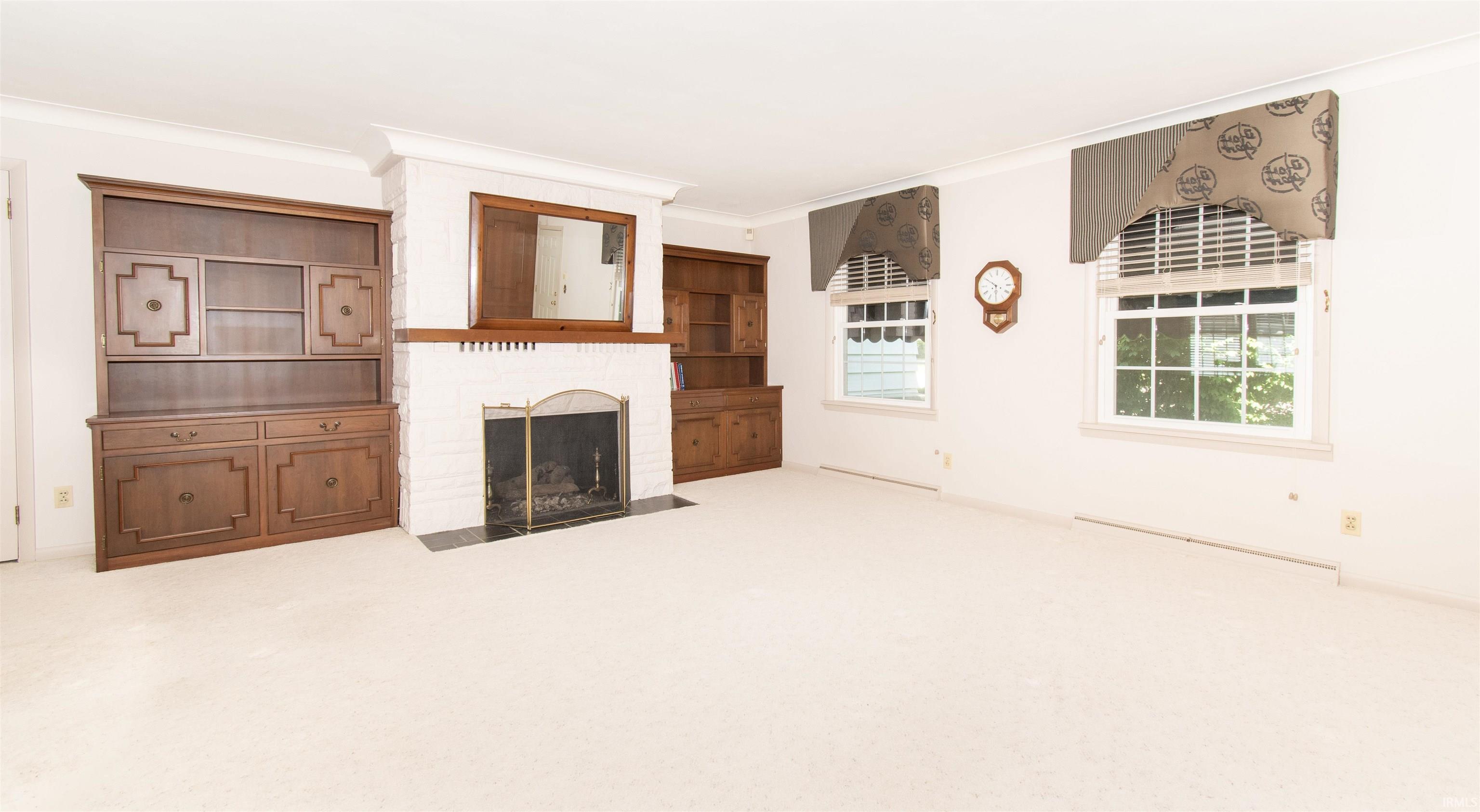 Spacious room featuring a white painted brick fireplace with a dark stone hearth, two built-in wood cabinetry units, and two windows with blinds and valances