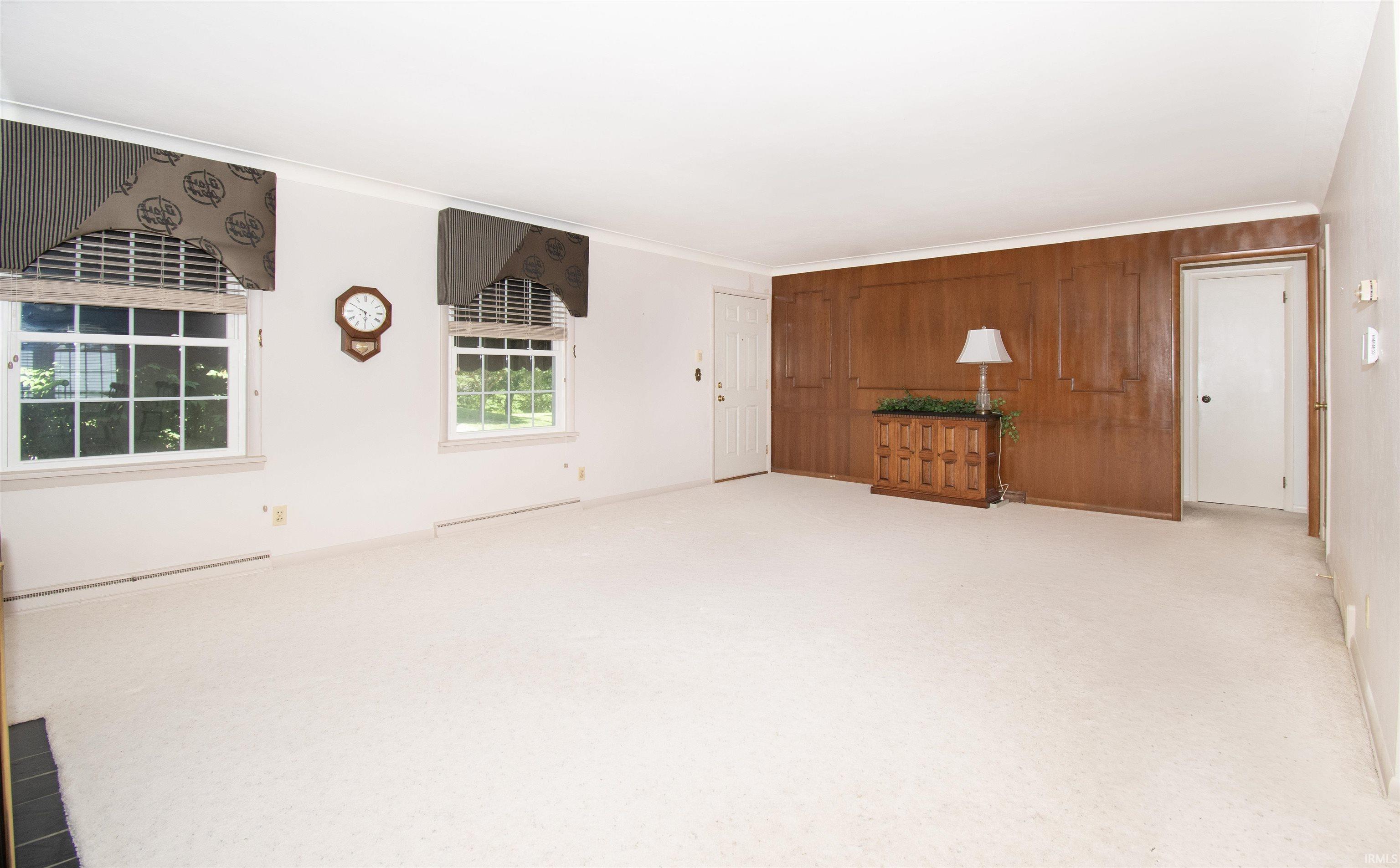 Spacious room featuring white carpeting, white walls, two double-hung windows with white frames, crown molding, and a paneled wood accent wall