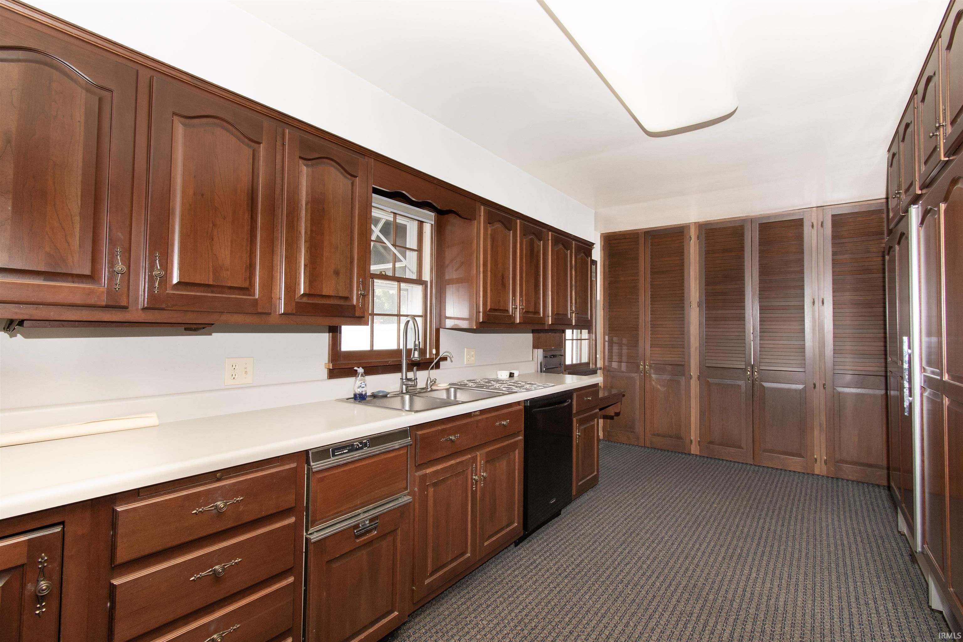 Kitchen featuring extensive wood cabinetry, white countertops, stainless steel sink with a gooseneck faucet, integrated dishwasher, and textured carpet flooring