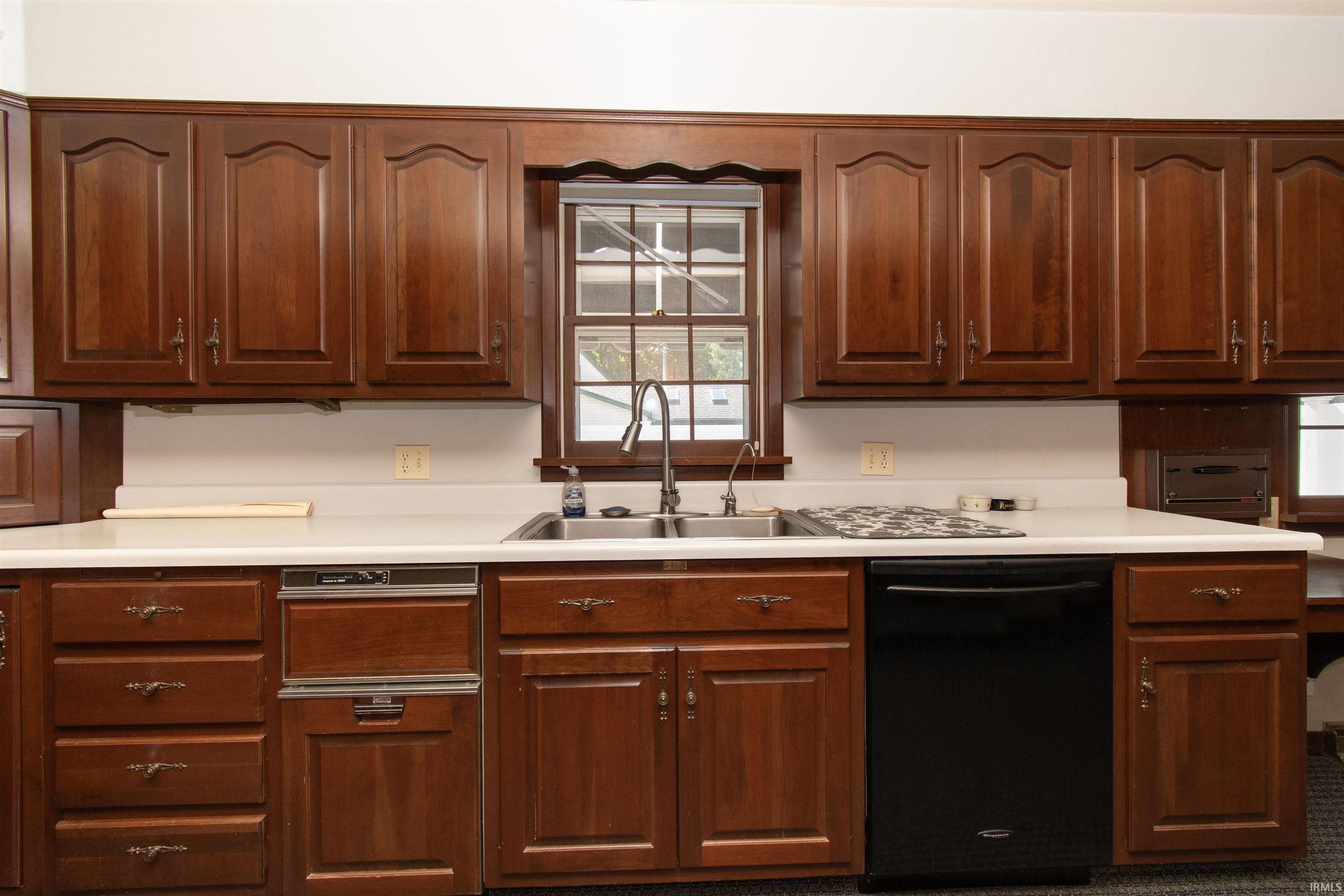Kitchen featuring extensive wood cabinetry with decorative hardware, a double basin sink with a gooseneck faucet, light-toned countertops, and a built-in black dishwasher