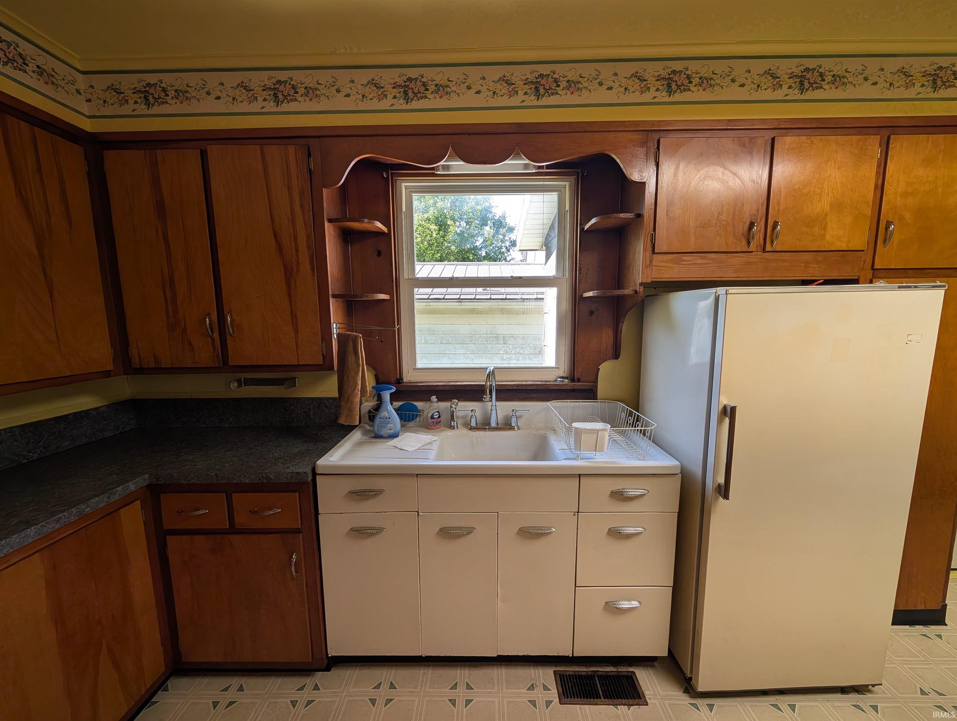 Kitchen featuring wood cabinetry, a white double-basin sink, an integrated window, a freestanding refrigerator, and patterned floor tiles