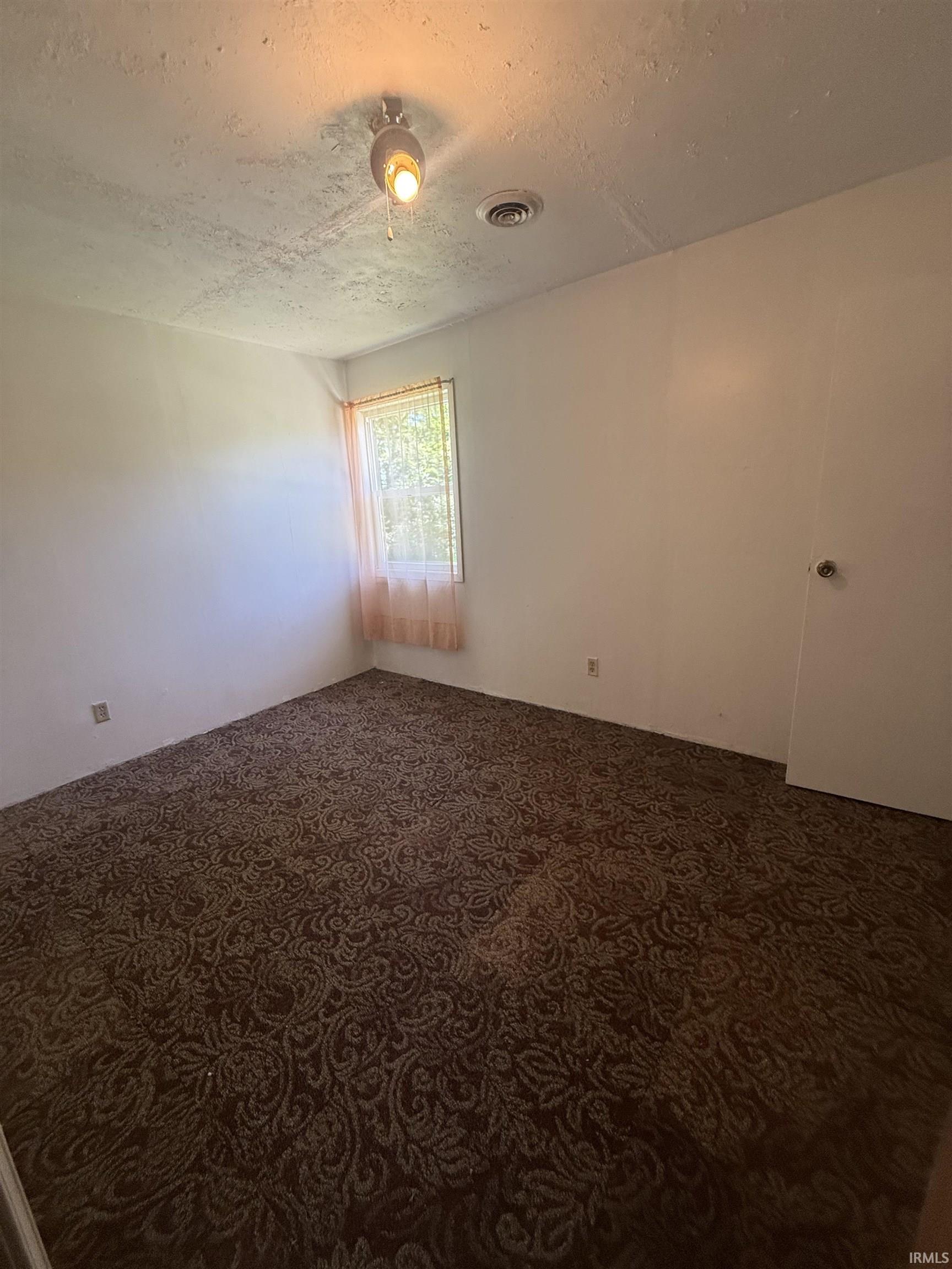 Room with patterned brown carpet flooring, white walls, and a single window with a sheer curtain