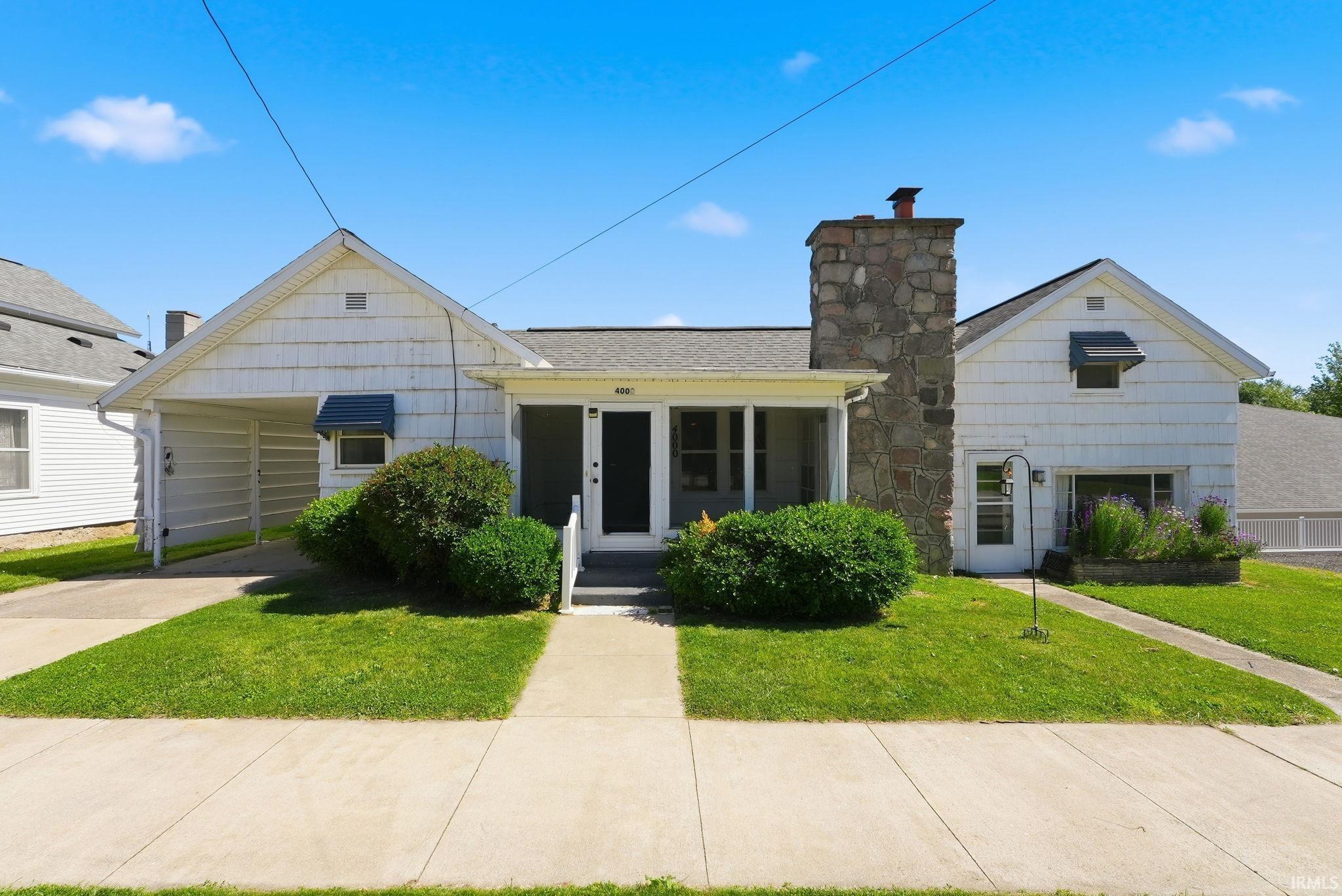 White horizontal siding exterior with a prominent stone chimney, covered porch, carport, and manicured lawn