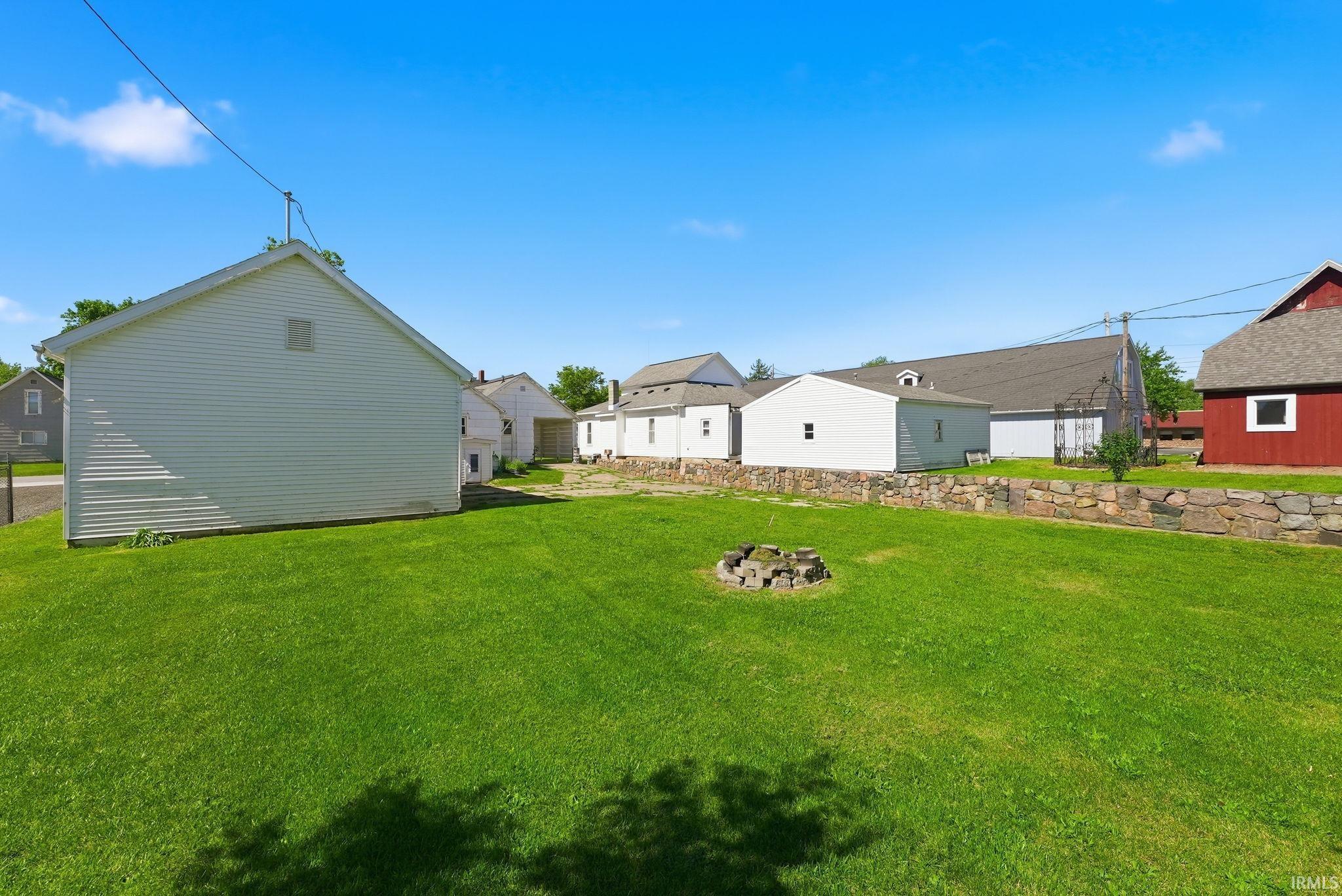 Expansive grassy yard area featuring a stone fire pit, a stone retaining wall, and multiple outbuildings with varied architectural styles