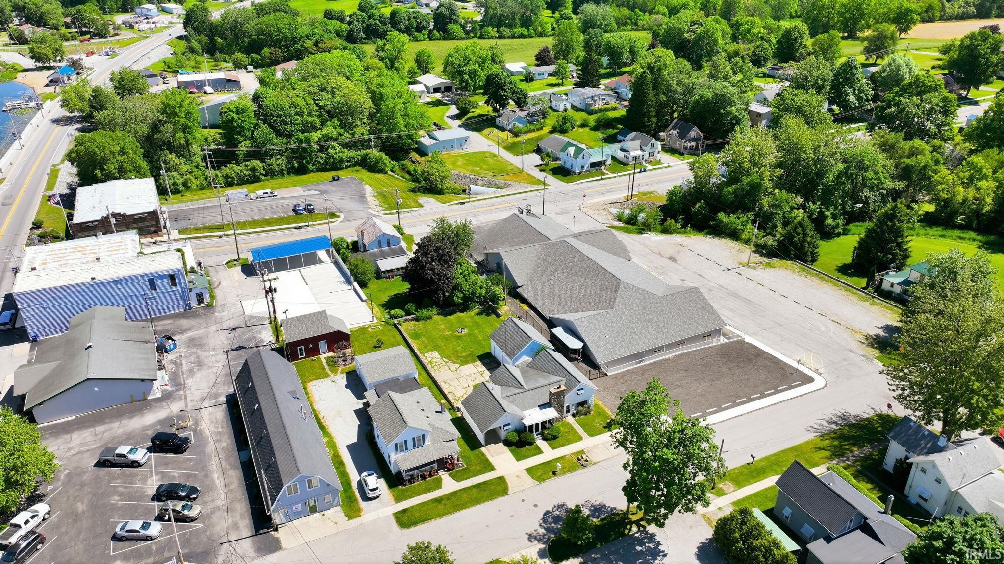 Aerial perspective highlighting commercial and residential properties, including a large building with a multi-gabled roof, multiple parking areas, and mature trees