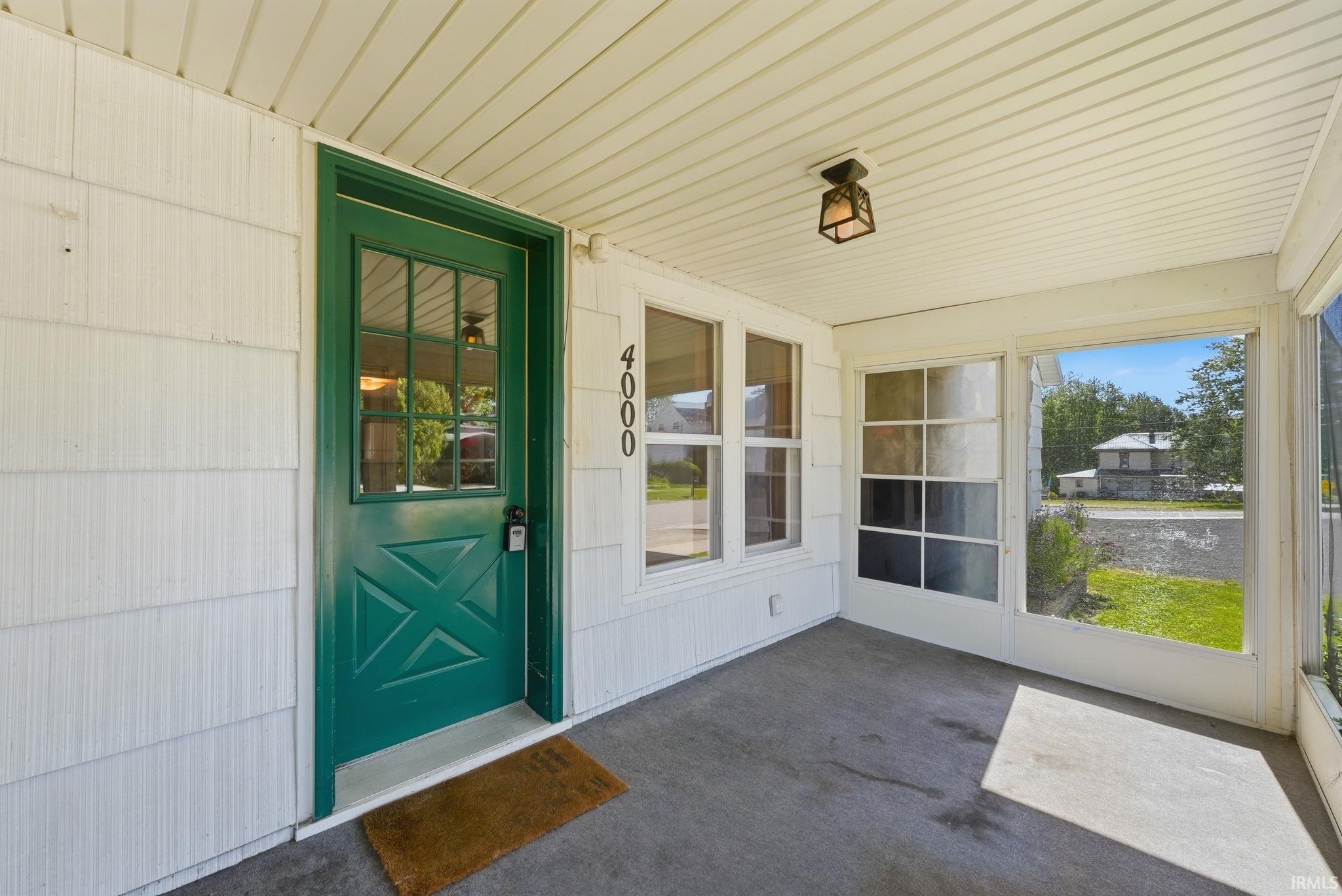 Enclosed front porch featuring a vibrant green entry door with glass panes, white siding, a textured concrete floor, and a classic flush-mount ceiling light