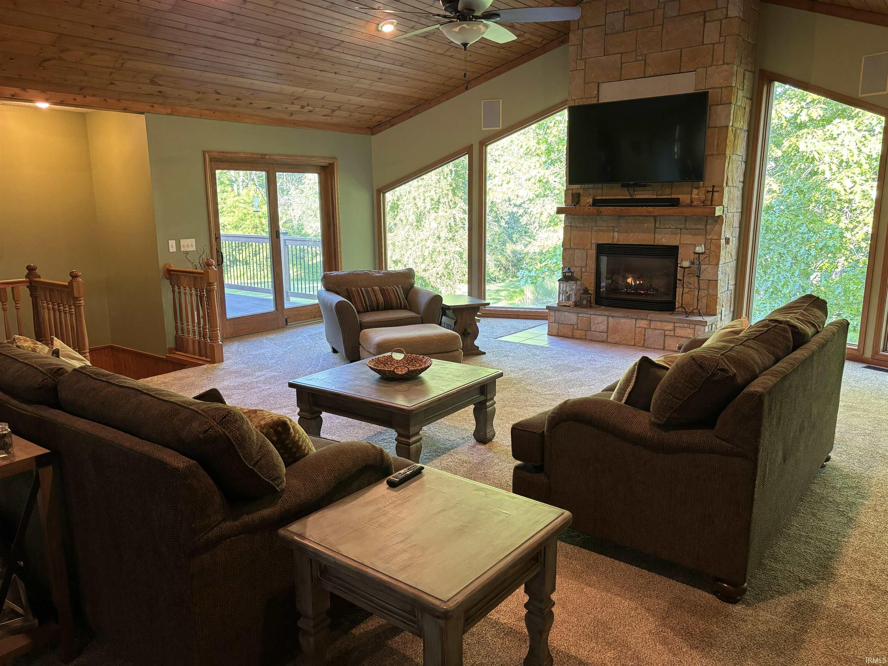 Living area featuring a stone-clad fireplace with a wood mantel, a wood-paneled vaulted ceiling with a ceiling fan, expansive windows, a sliding glass door leading to a balcony, and carpet flooring