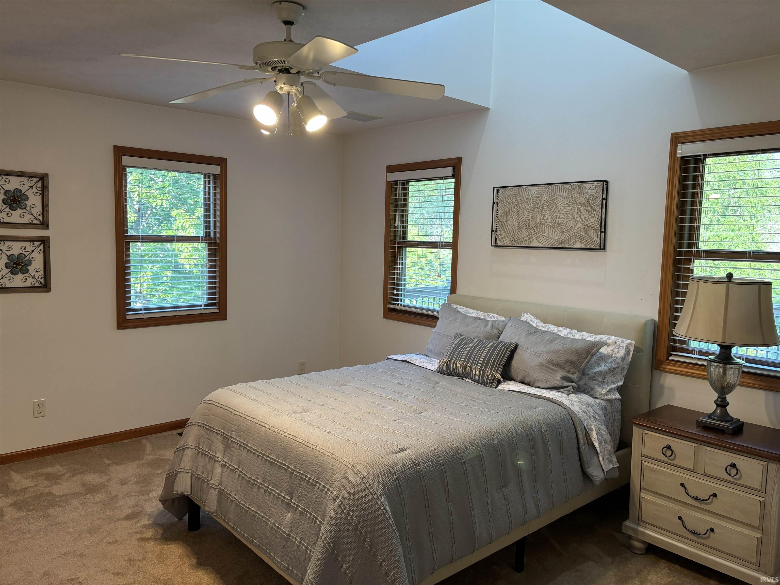 Carpeted room featuring a ceiling fan with integrated lighting, two windows with wood-trim framing, and a vaulted ceiling with a skylight