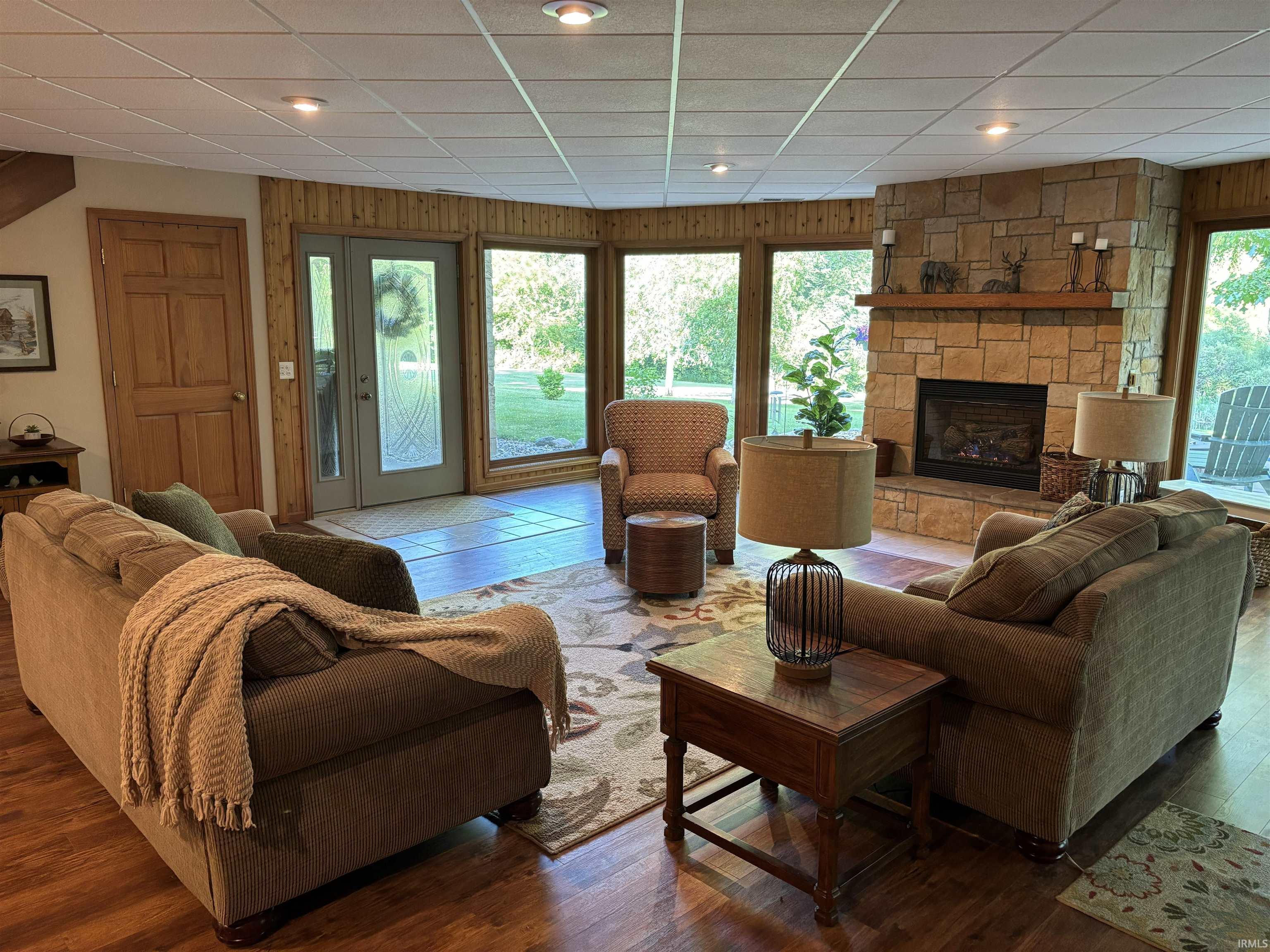 Spacious living area featuring a stone-clad fireplace with a wood mantel, wood-finish flooring, and extensive natural light from multiple windows