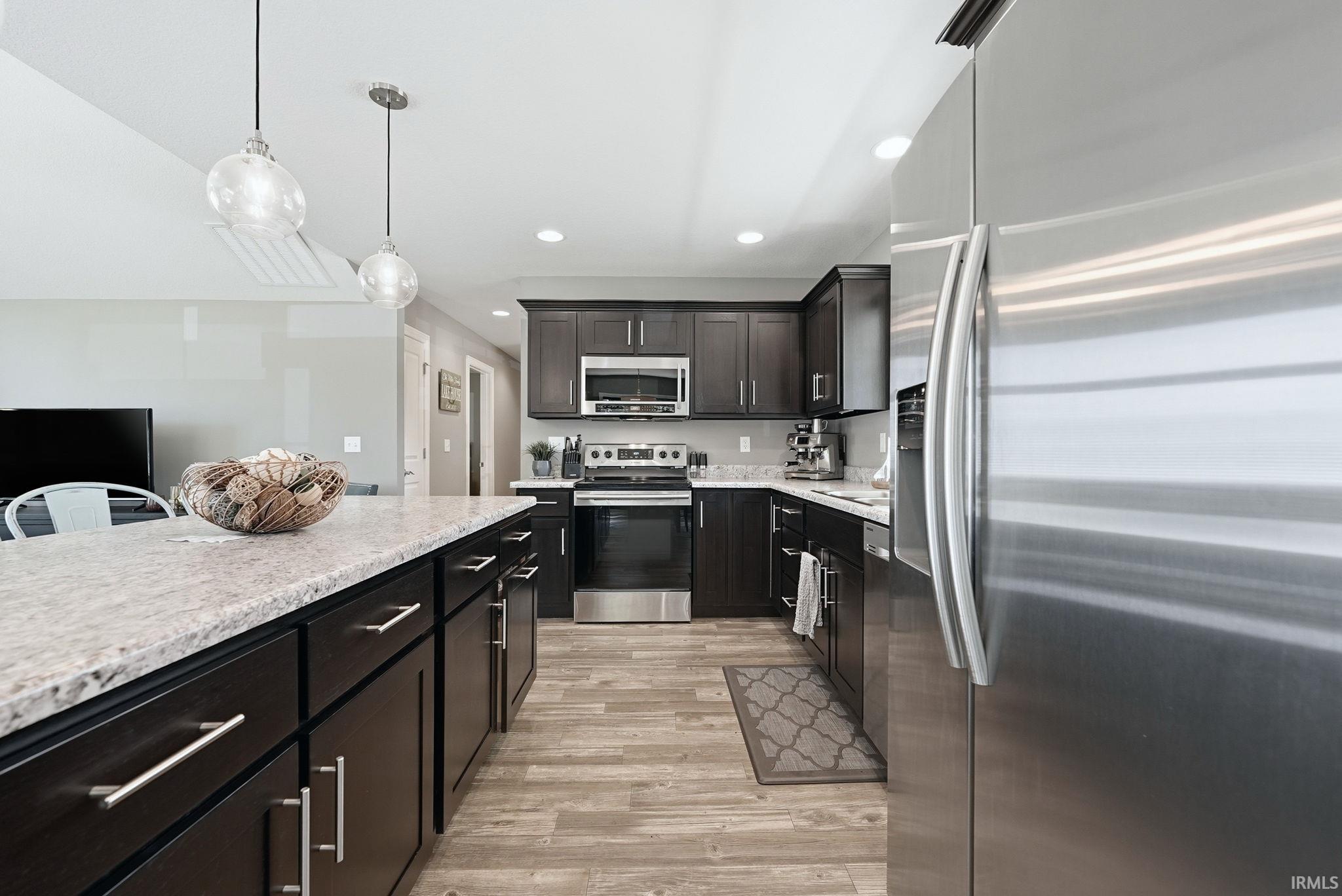 Kitchen featuring a spacious island with a light-toned countertop, dark wood-finish cabinetry, stainless steel appliances, wood-finish flooring, and recessed lighting