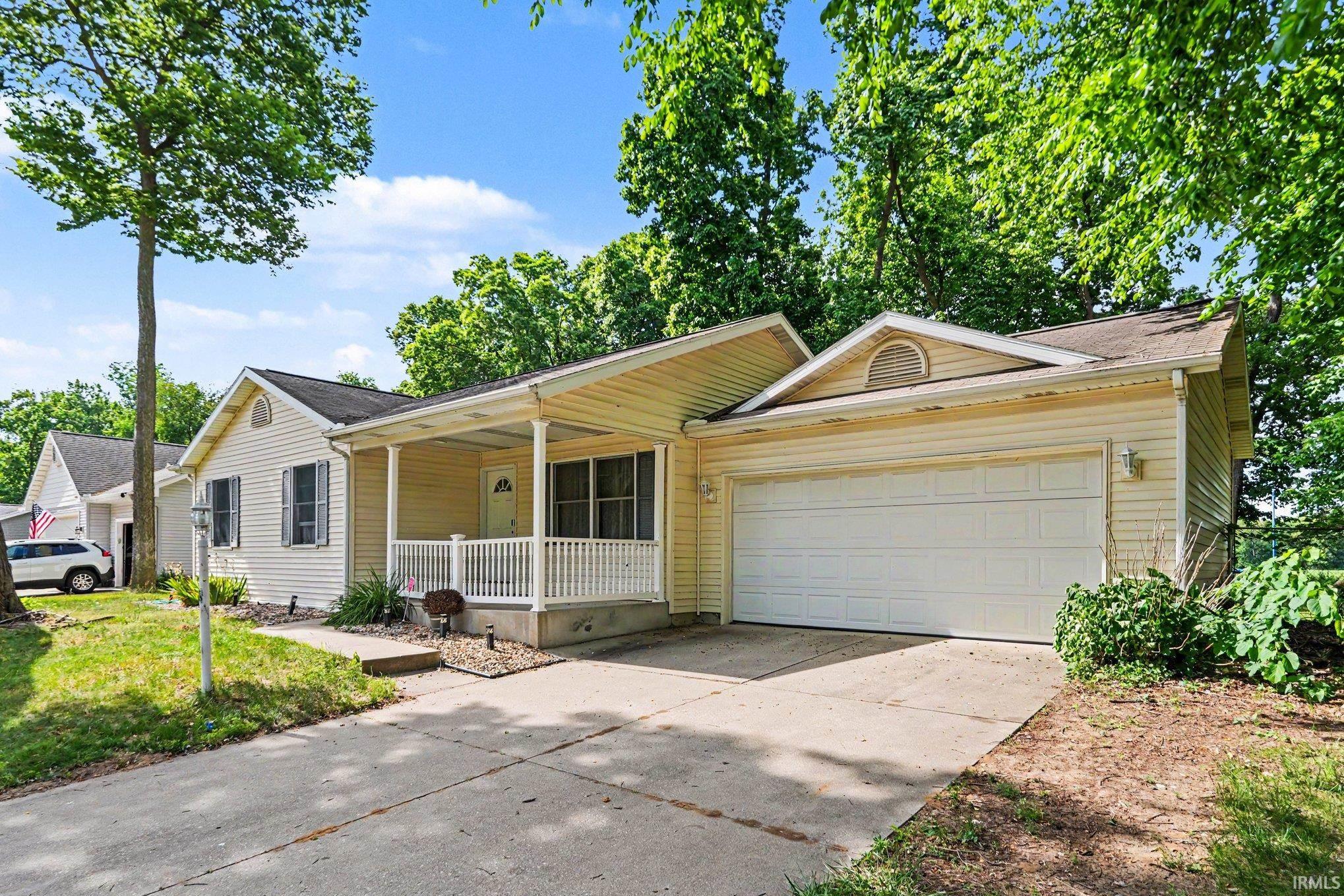 Ranch-style exterior with light-colored siding, covered front porch, attached garage, paved driveway, and mature landscaping