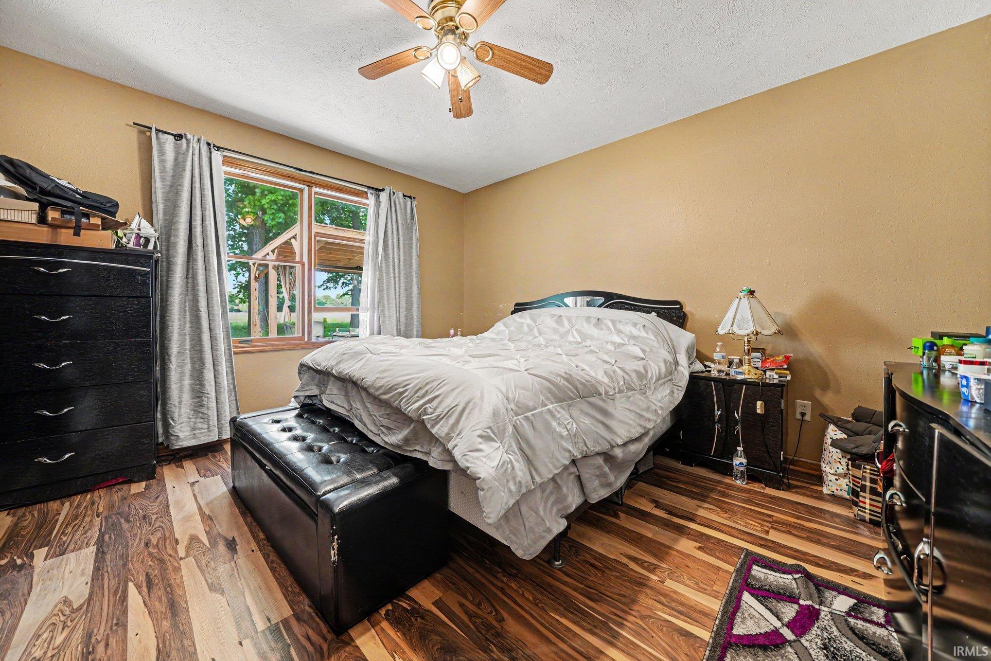 Bedroom featuring wood-finish flooring, a window with exterior views, light tan walls, and a ceiling fan with integrated lighting
